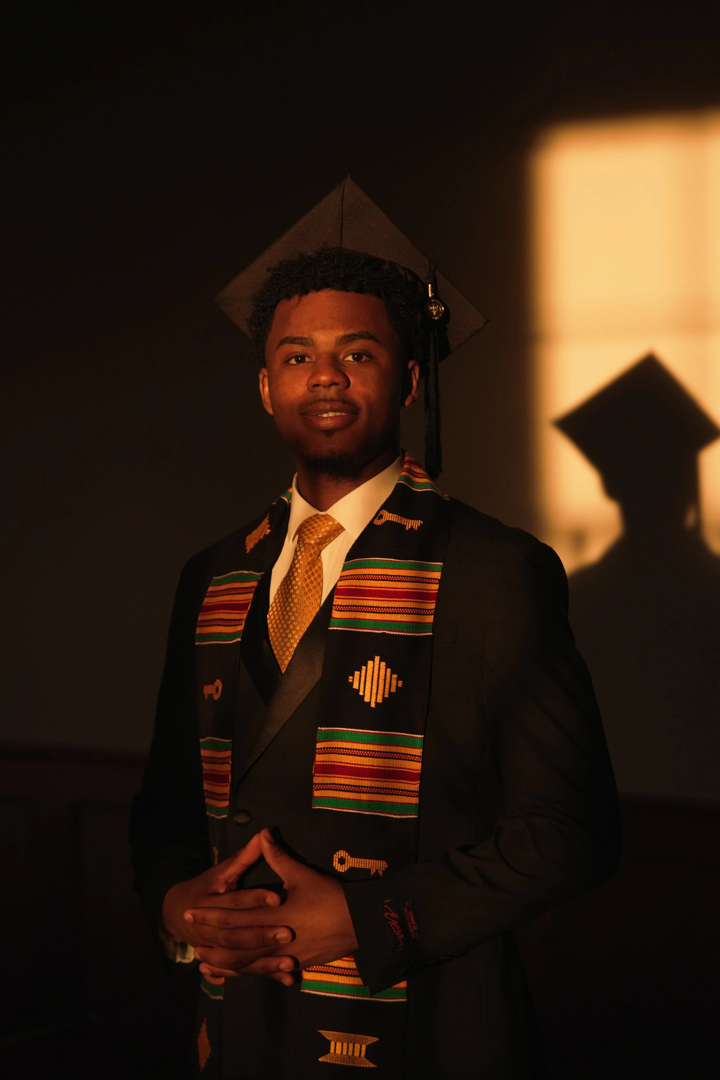 A young man in a graduation cap and gown, wearing a colorful kente cloth stole, standing against a dark background with a glowing light on the right side.