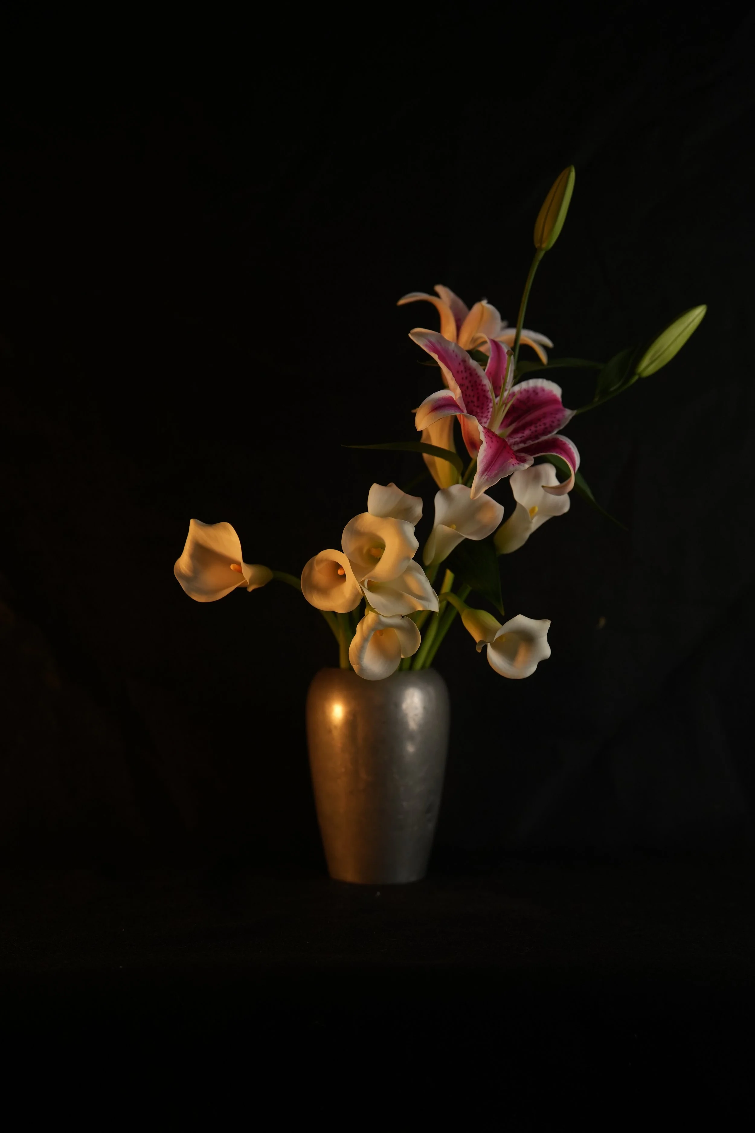 A bouquet of lilies in a silver vase against a dark background.