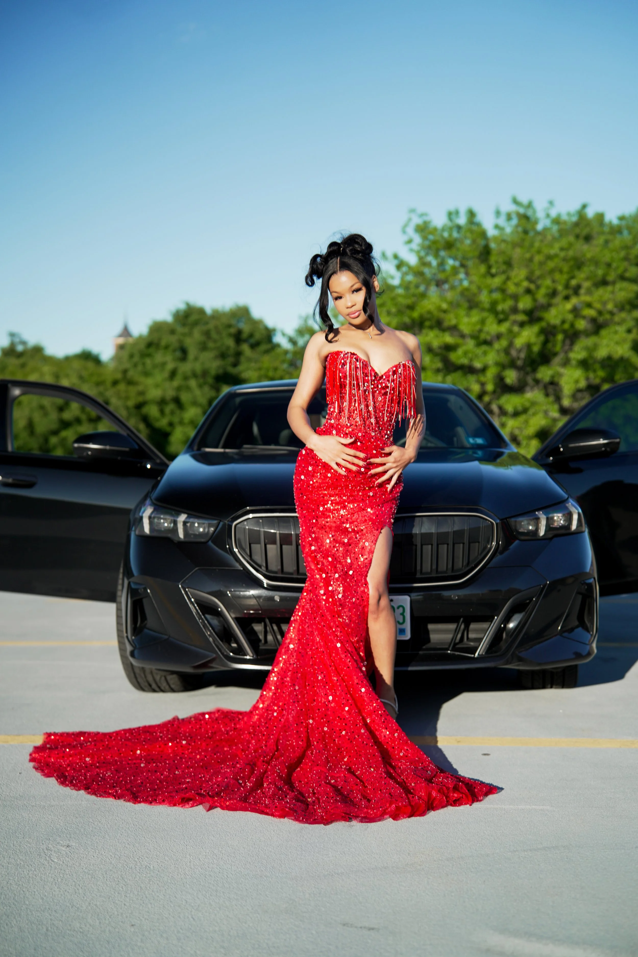 Woman in a red, sequined evening gown standing in front of a black sports car in a parking lot with trees and blue sky in the background.