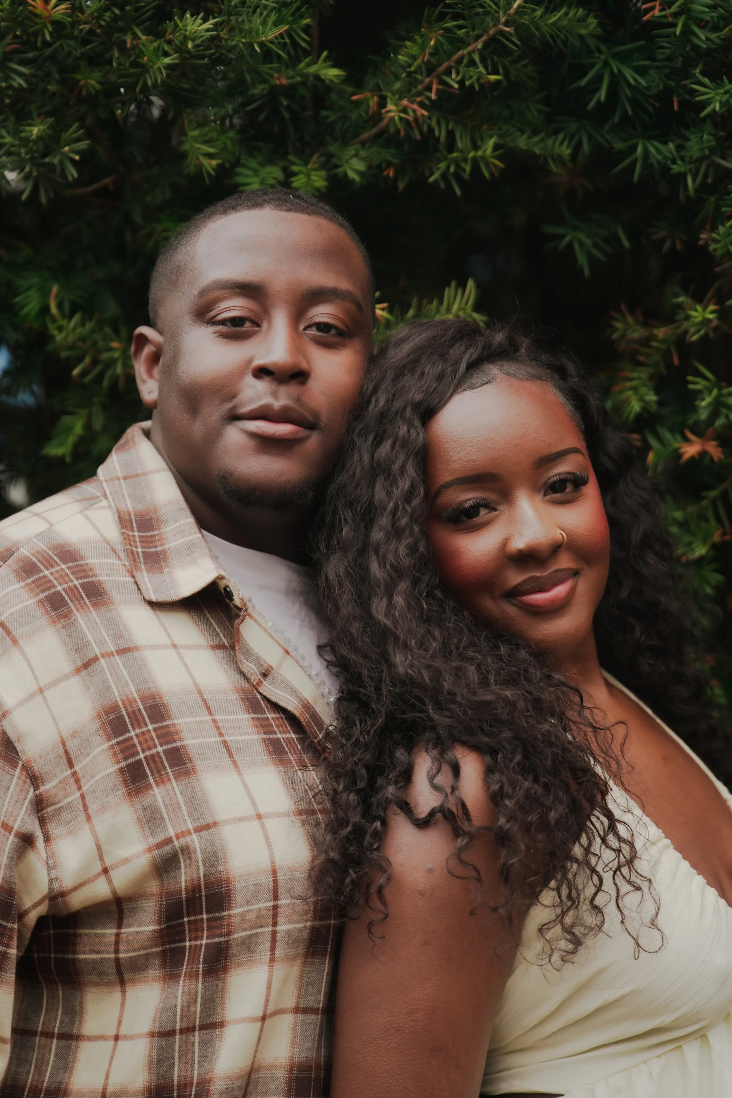 A close-up of a young couple standing outdoors. The man has short hair and is wearing a plaid shirt, while the woman has long, curly hair and is wearing a light-colored top. They are smiling slightly and are positioned close to each other with lush g