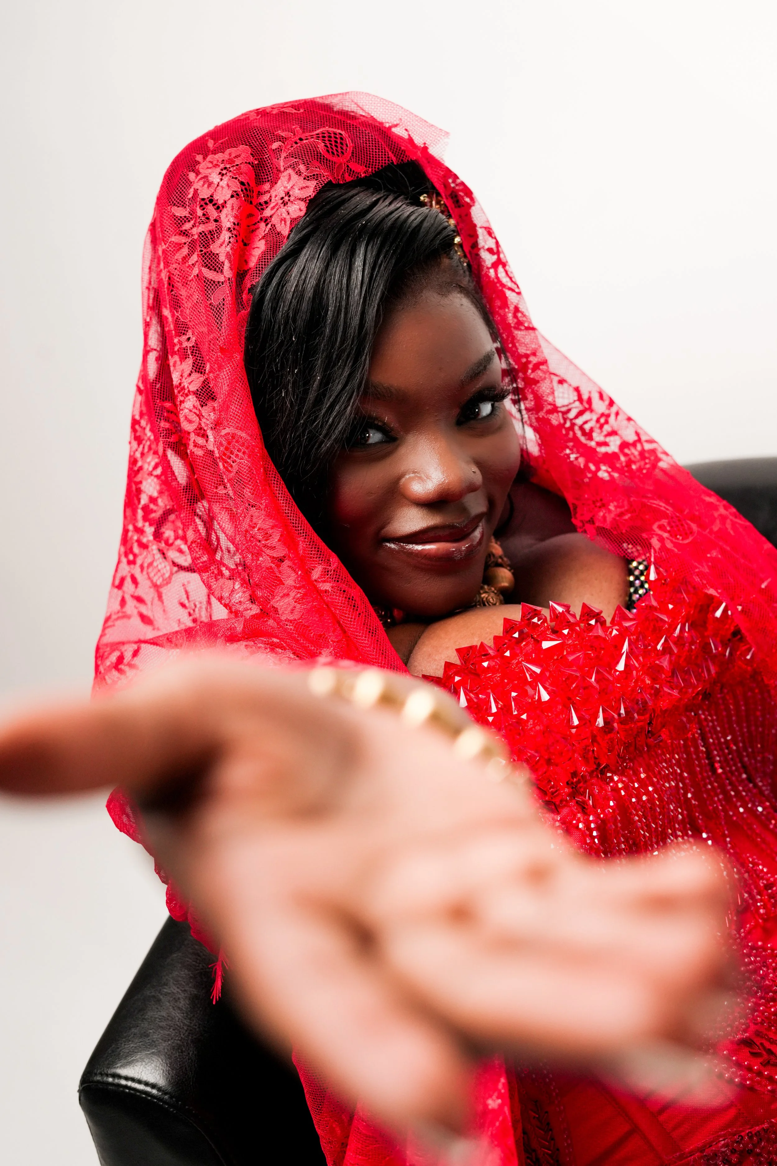 A woman with dark braided hair, wearing a red lace veil and matching red dress with embellishments, sitting on a black chair against a plain white background.