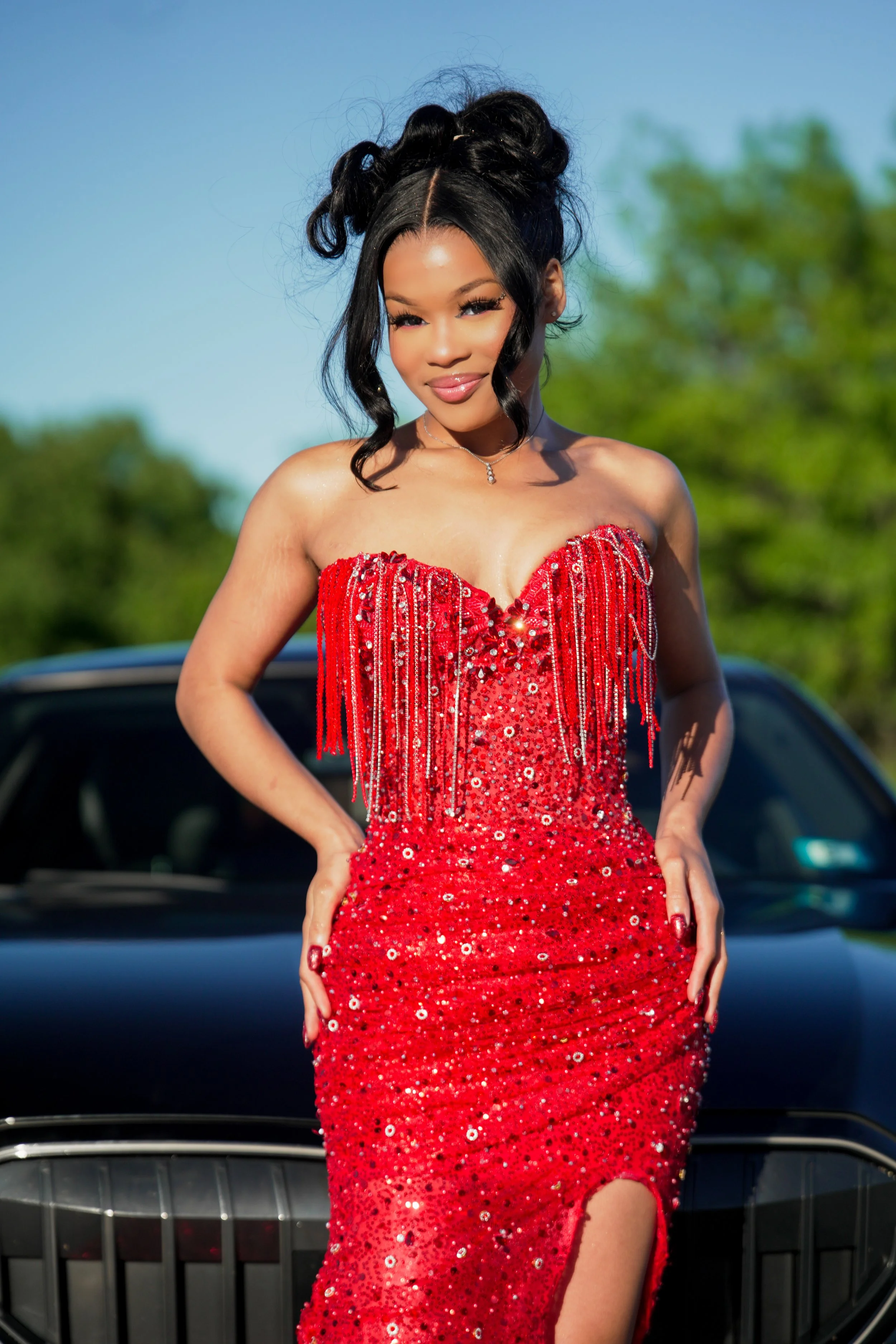 A woman in a strapless red sequined dress with a thigh-high slit posing outdoors on a sunny day, with a car visible behind her and greenery in the background.