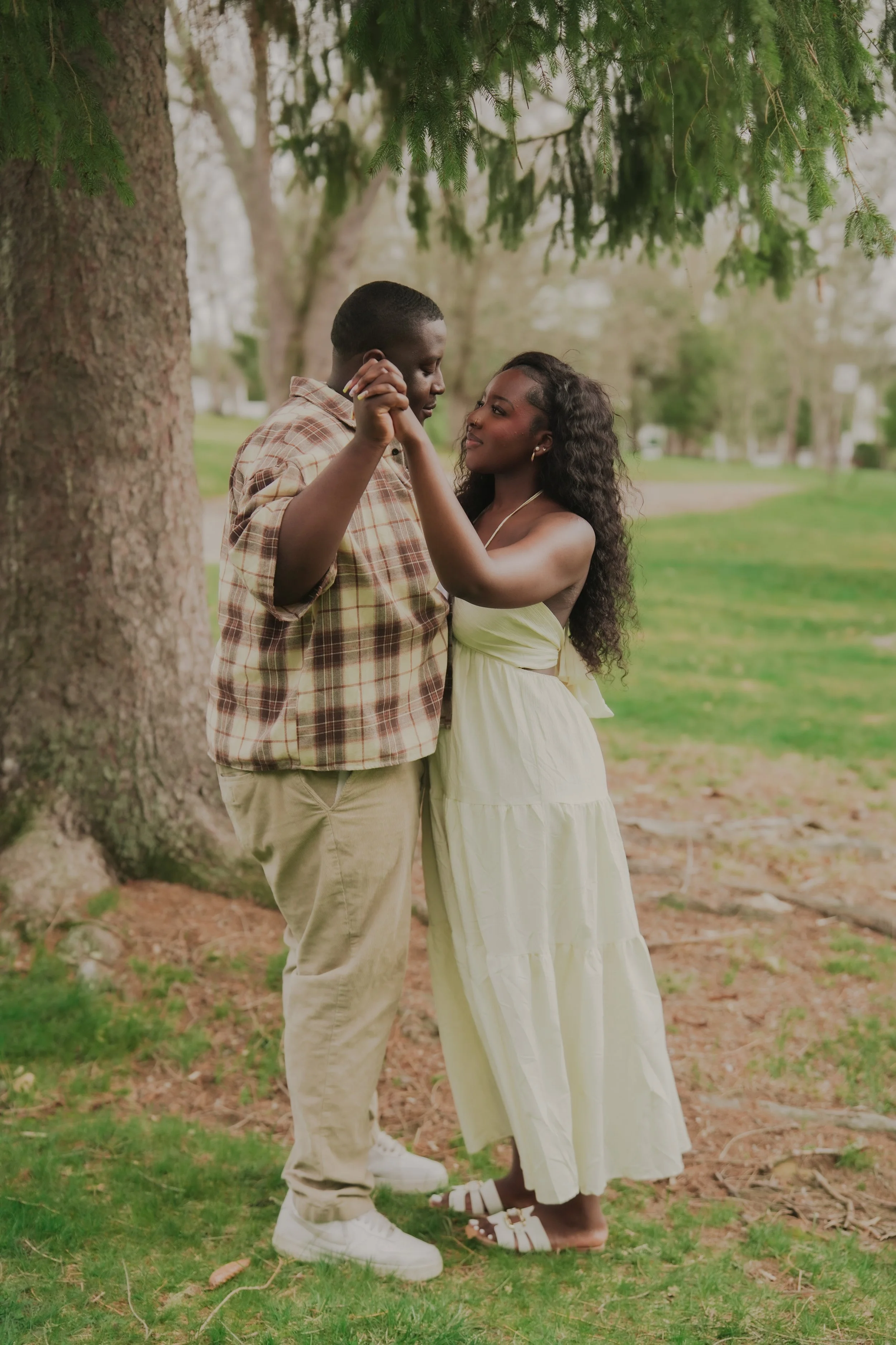 A young couple dancing outdoors under a large tree in a park. The man is wearing a plaid shirt and beige pants, and the woman is dressed in a white summer dress and sandals. They gaze into each other's eyes, holding hands, with a green park and trees