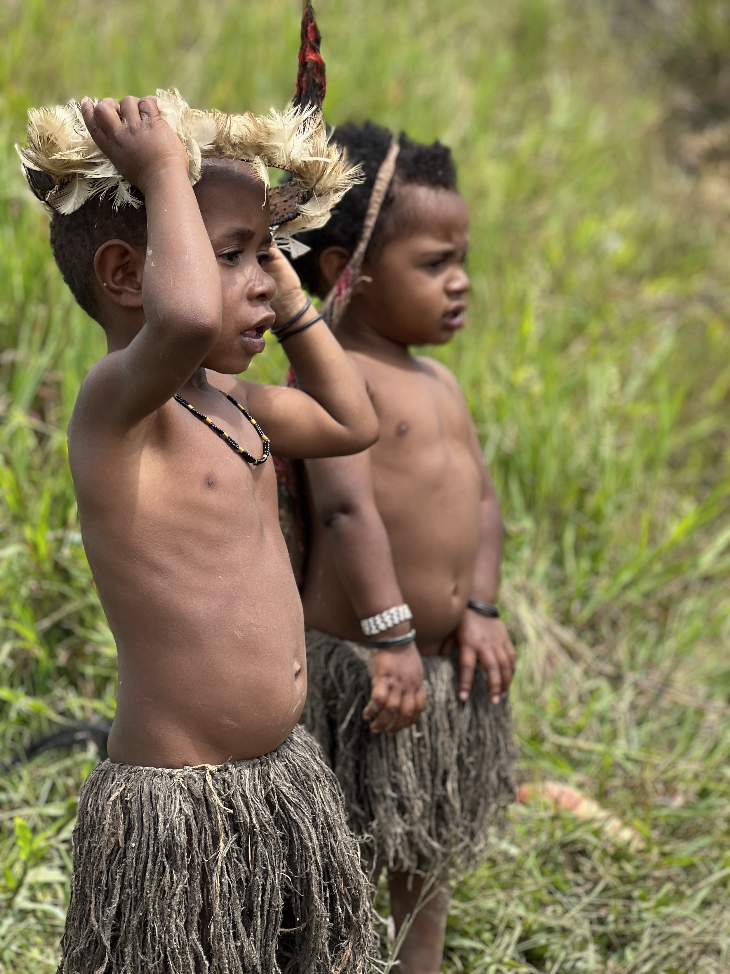Two young boys from an indigenous tribe wearing traditional grass skirts, standing in a grassy outdoor area, with one adjusting a feathered headdress.