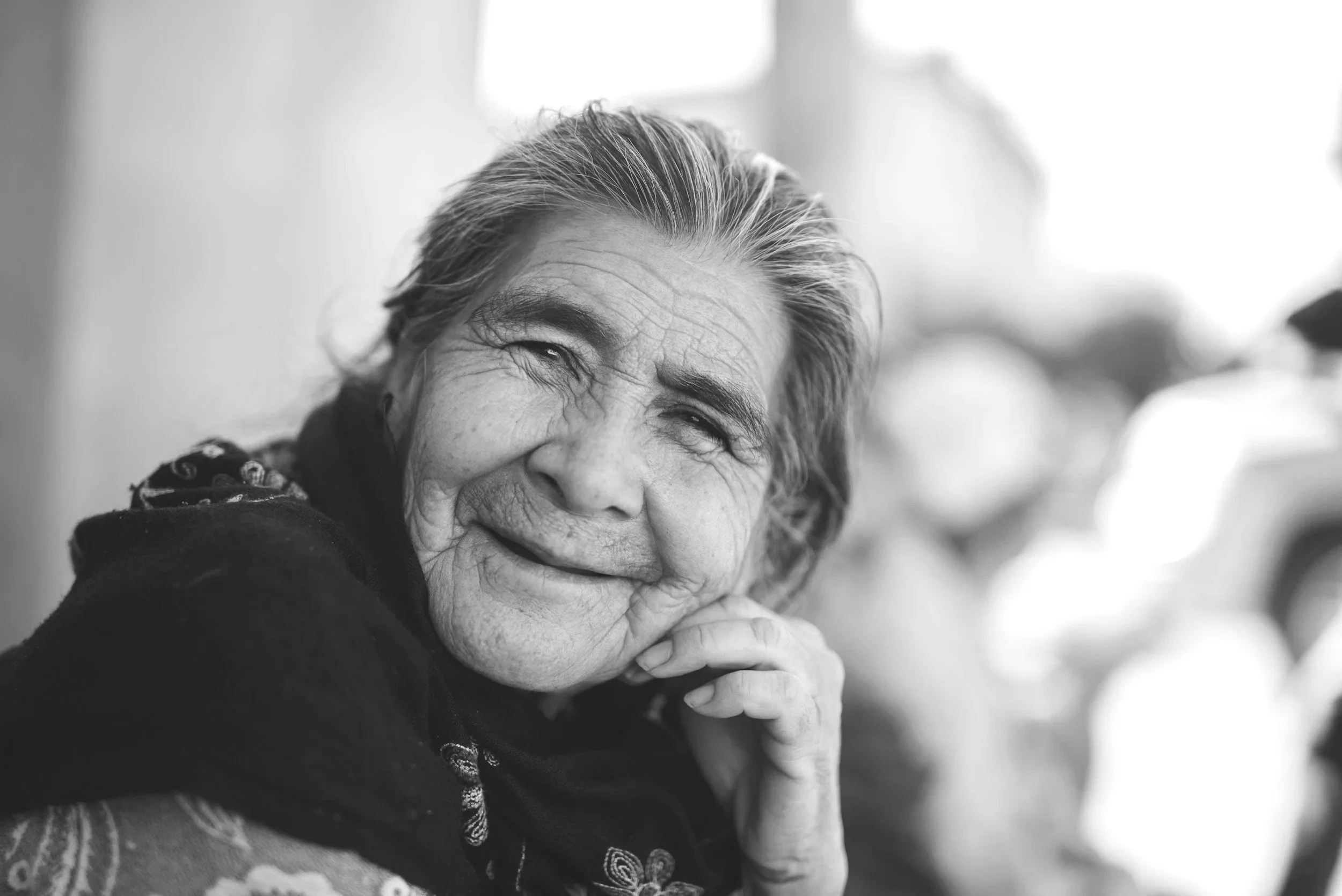 Black and white photo of an elderly woman smiling, with her hand resting on her chin, wearing a scarf.