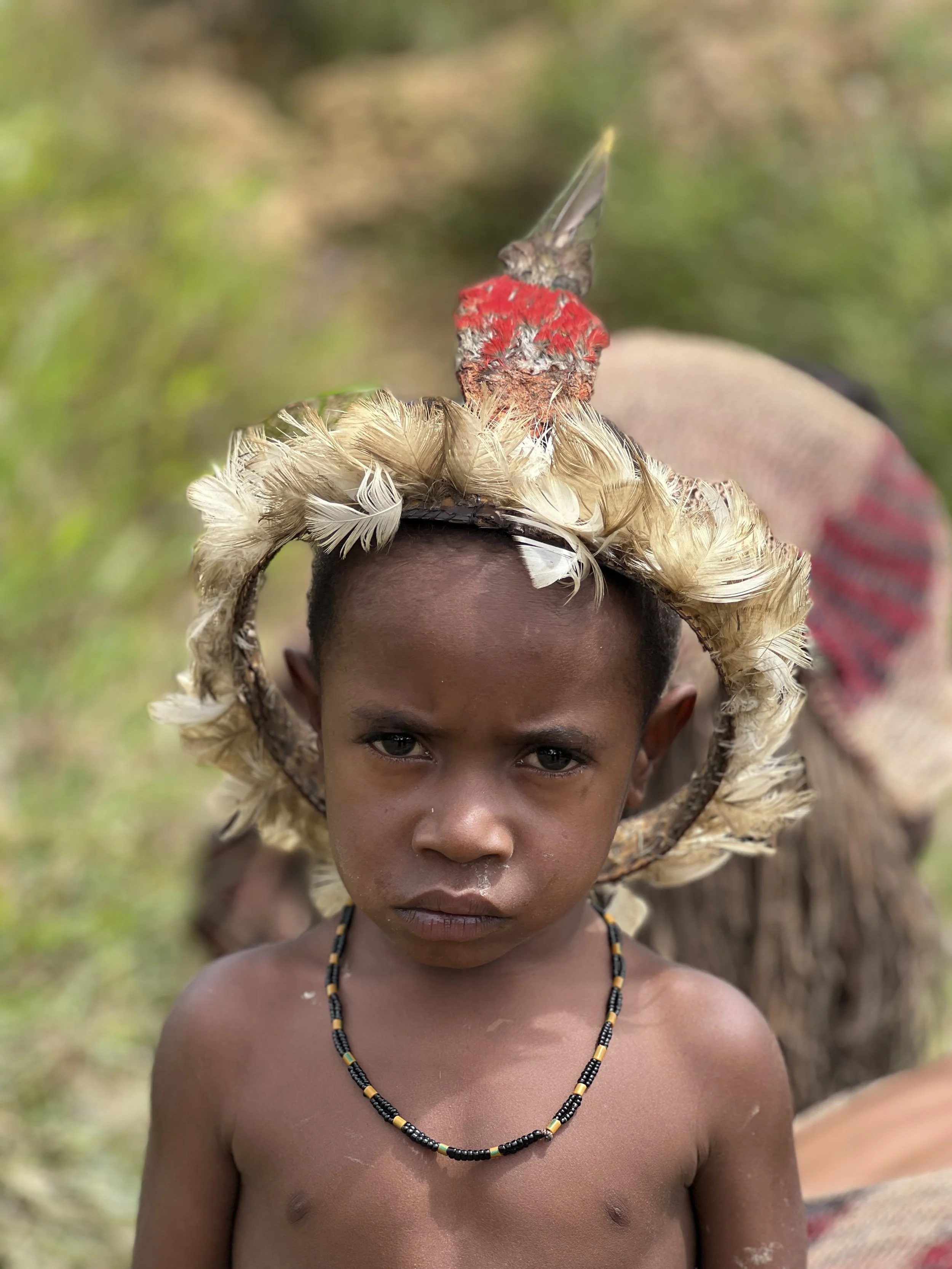 A young boy with a serious expression, wearing a beaded necklace, standing outdoors with a large hat decorated with feathers, topped with a small bird.