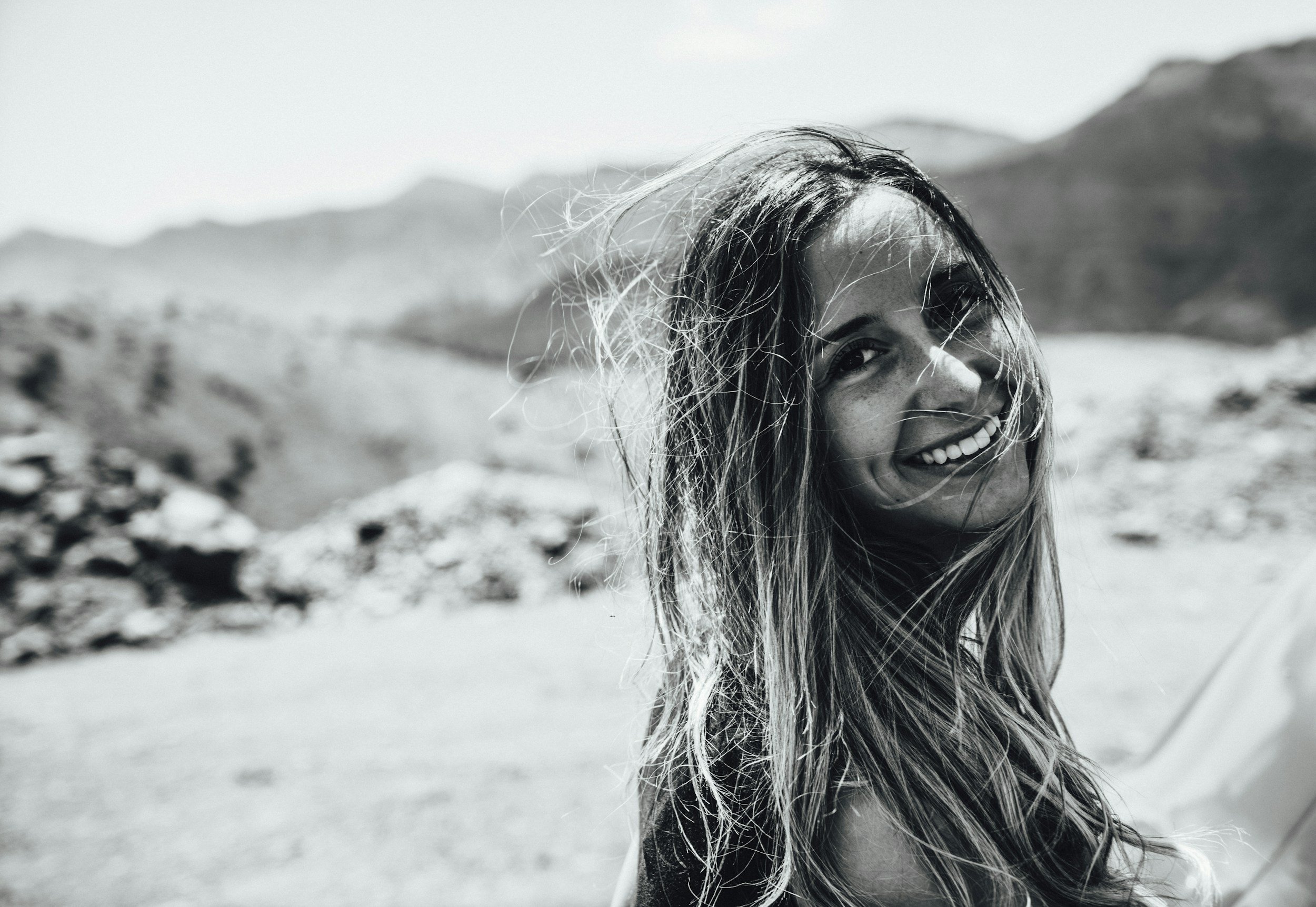 A smiling woman with long hair standing outdoors in a mountainous environment with rocky terrain in the background.