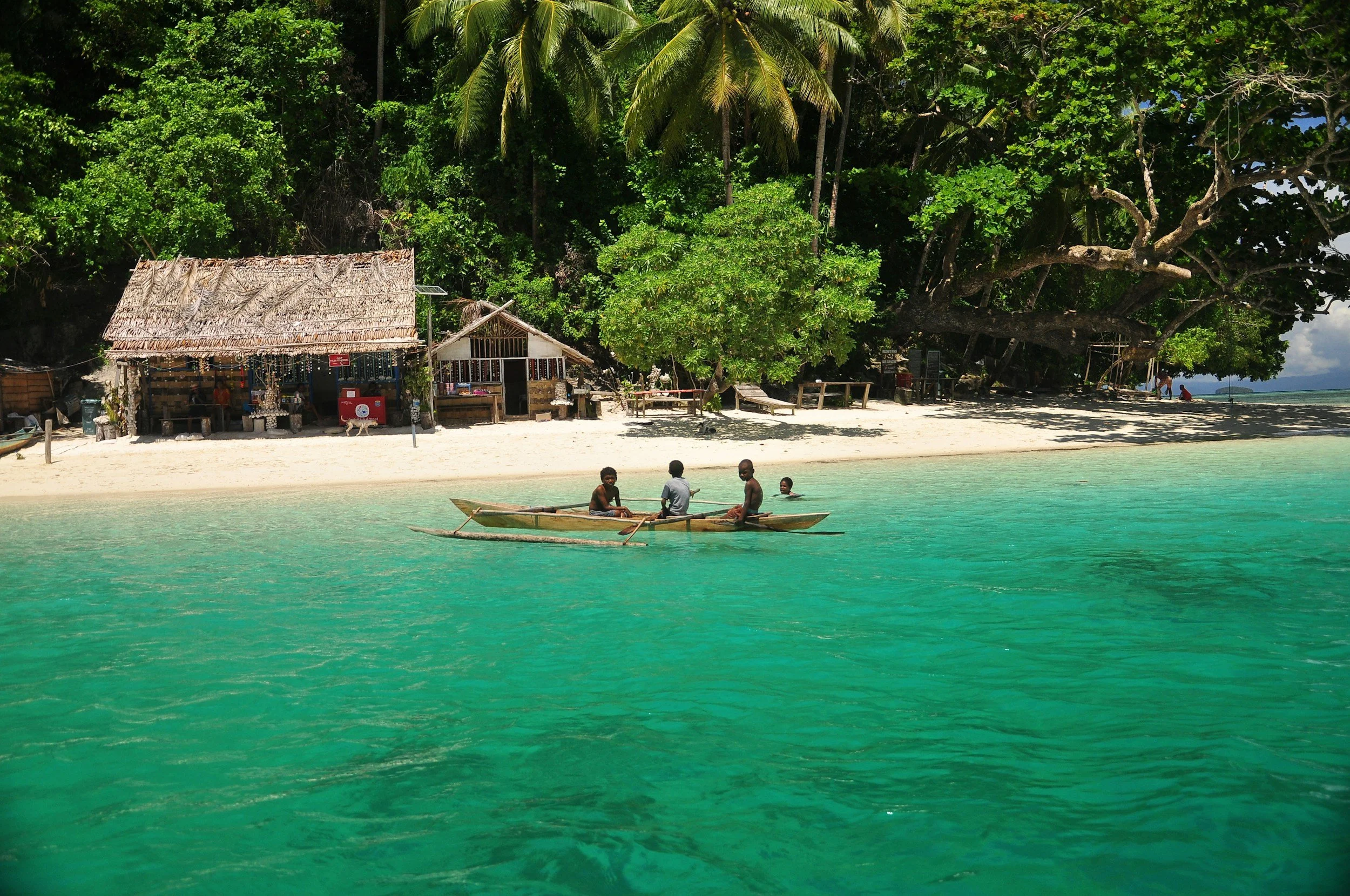 Four people in a small boat on turquoise water near a sandy beach with lush green trees and rustic huts in the background.