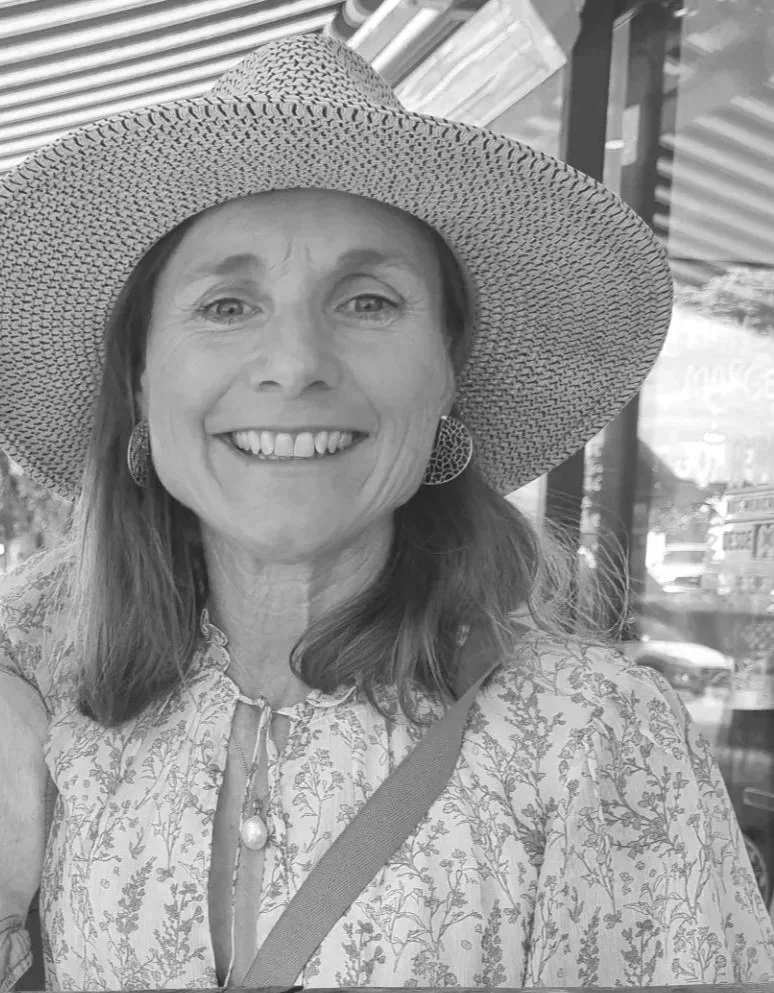A woman smiling, wearing a wide-brimmed hat, earrings, a floral patterned blouse, with a strap across her chest, standing outdoors with a building and trees in the background.