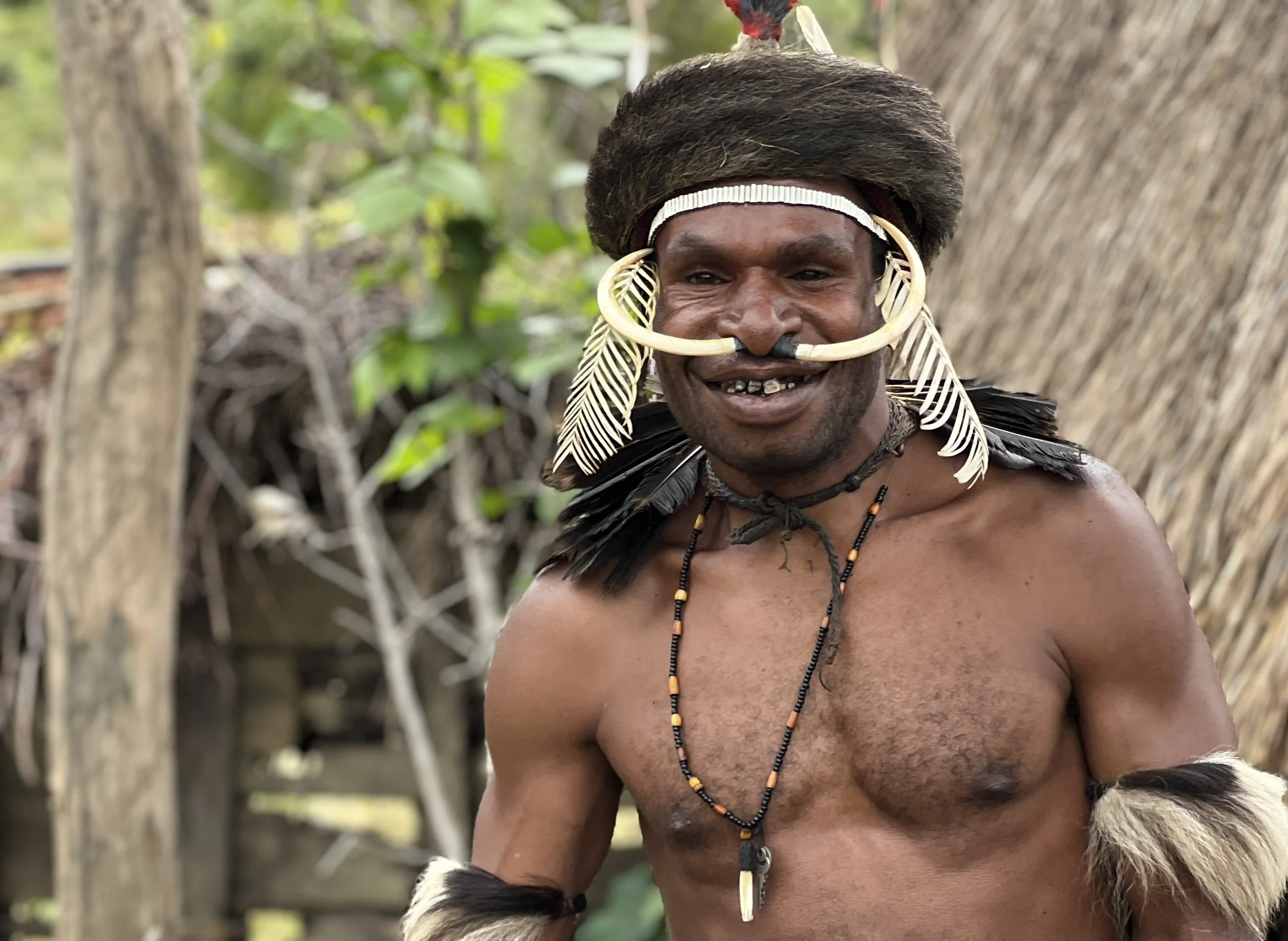A smiling man with traditional tribal accessories, including a headpiece with carved decorations, feathered shoulder adornments, and a necklace, standing outdoors near trees.