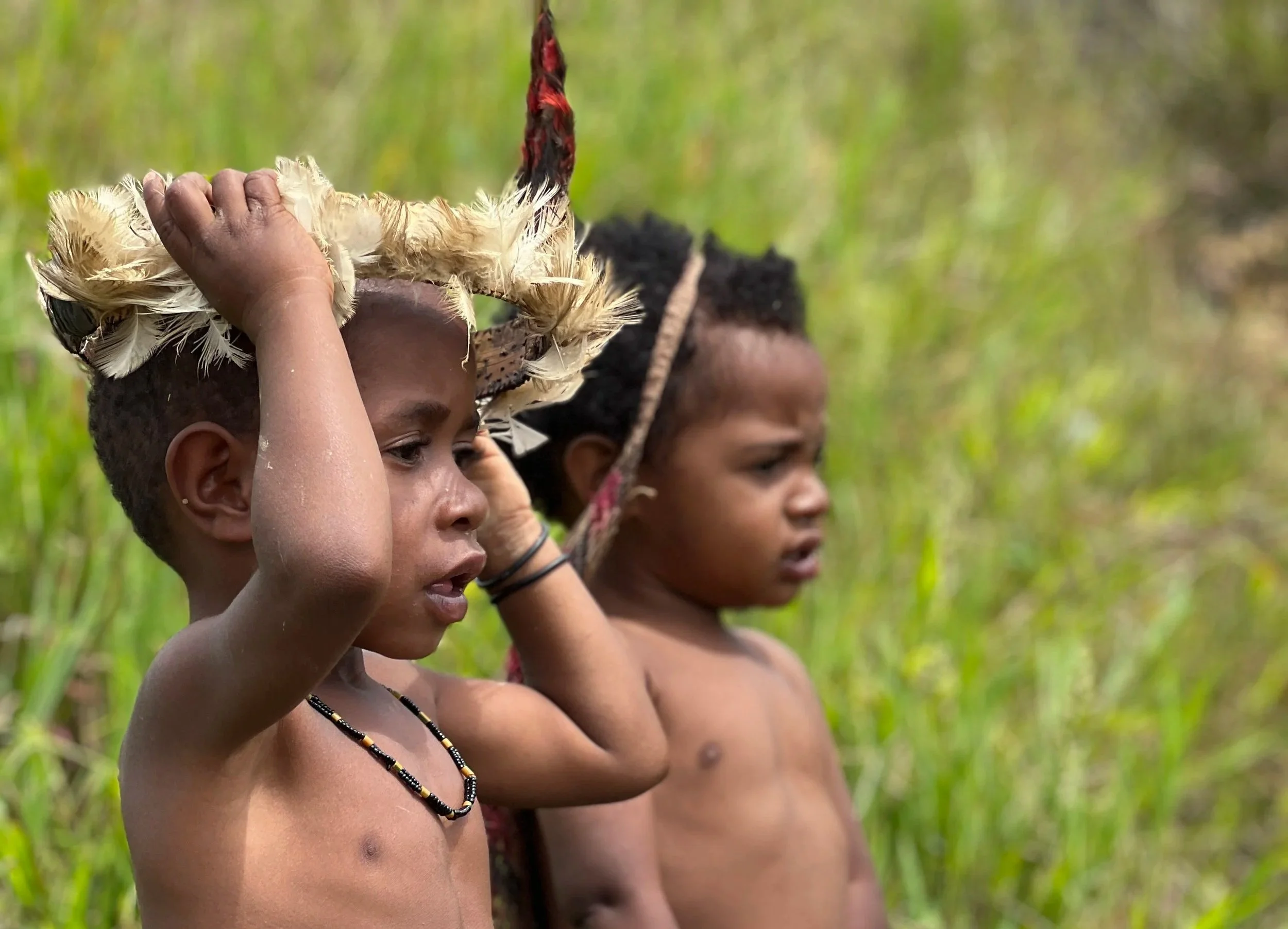 Two young children with dark skin and short curly hair, shirtless, standing in tall green grass, wearing traditional headgear and necklaces, with focused expressions.