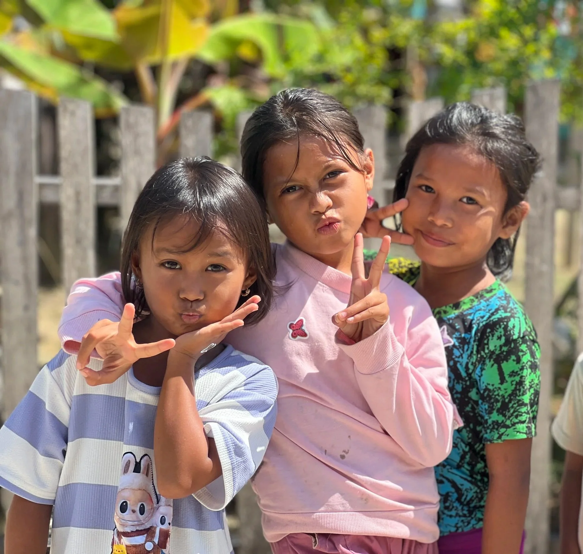 Three children standing outdoors, making playful facial expressions and hand gestures, in front of a wooden fence and green foliage.