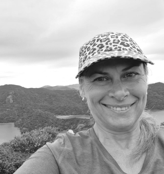 A woman smiling, wearing a leopard print cap, outdoors with a view of lakes and hills in the background.