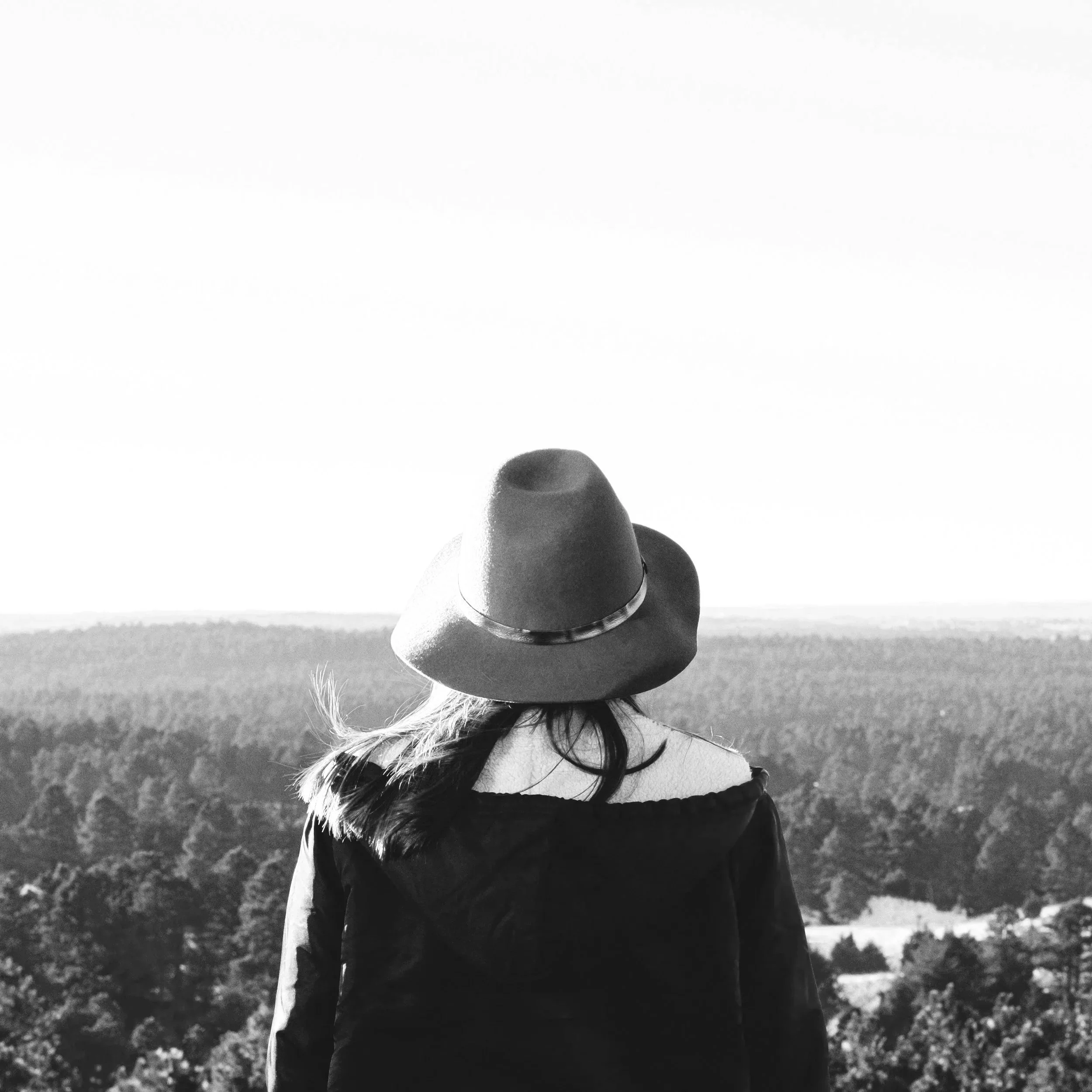 A woman with long hair wearing a wide-brimmed hat and coat stands facing a vast forested landscape in black and white.