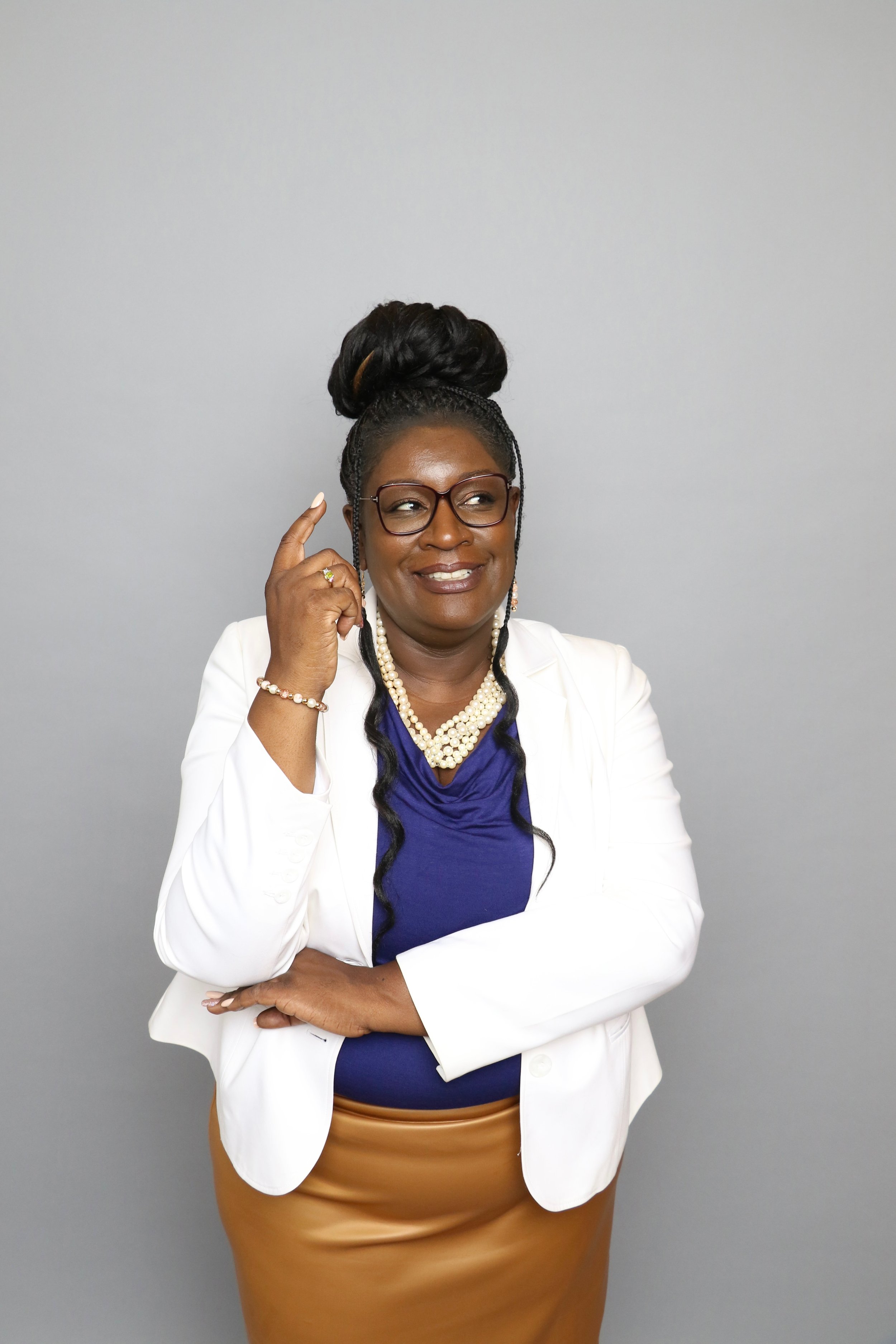 A confident woman with glasses, styled hair in an updo, wearing a white blazer over a blue top, a tan skirt, and pearl jewelry, standing against a plain grey background, smiling thoughtfully with her right hand touching her temple.