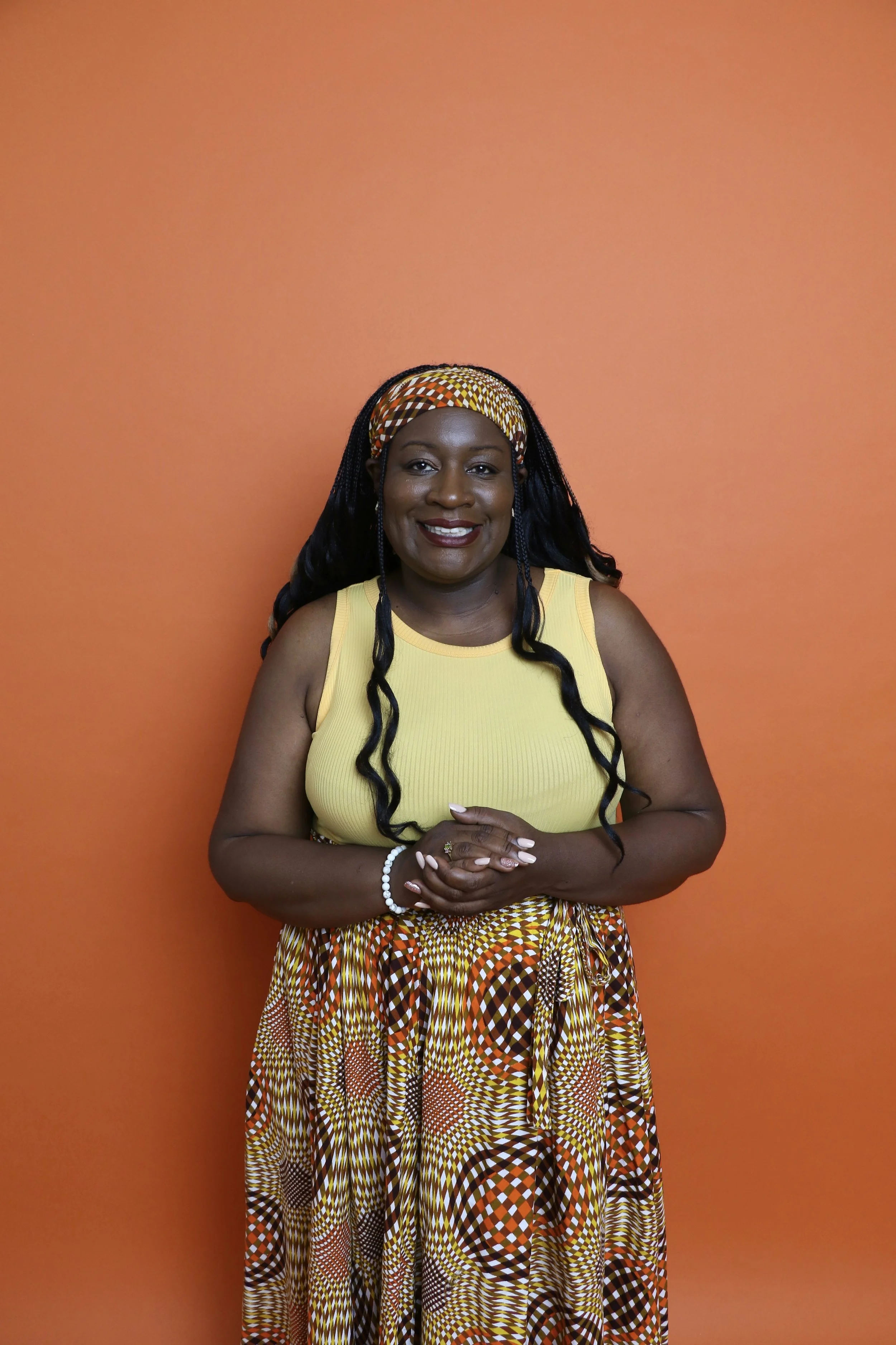 A woman wearing a yellow sleeveless top, patterned long skirt, and headscarf, standing against an orange background, smiling with hands clasped.
