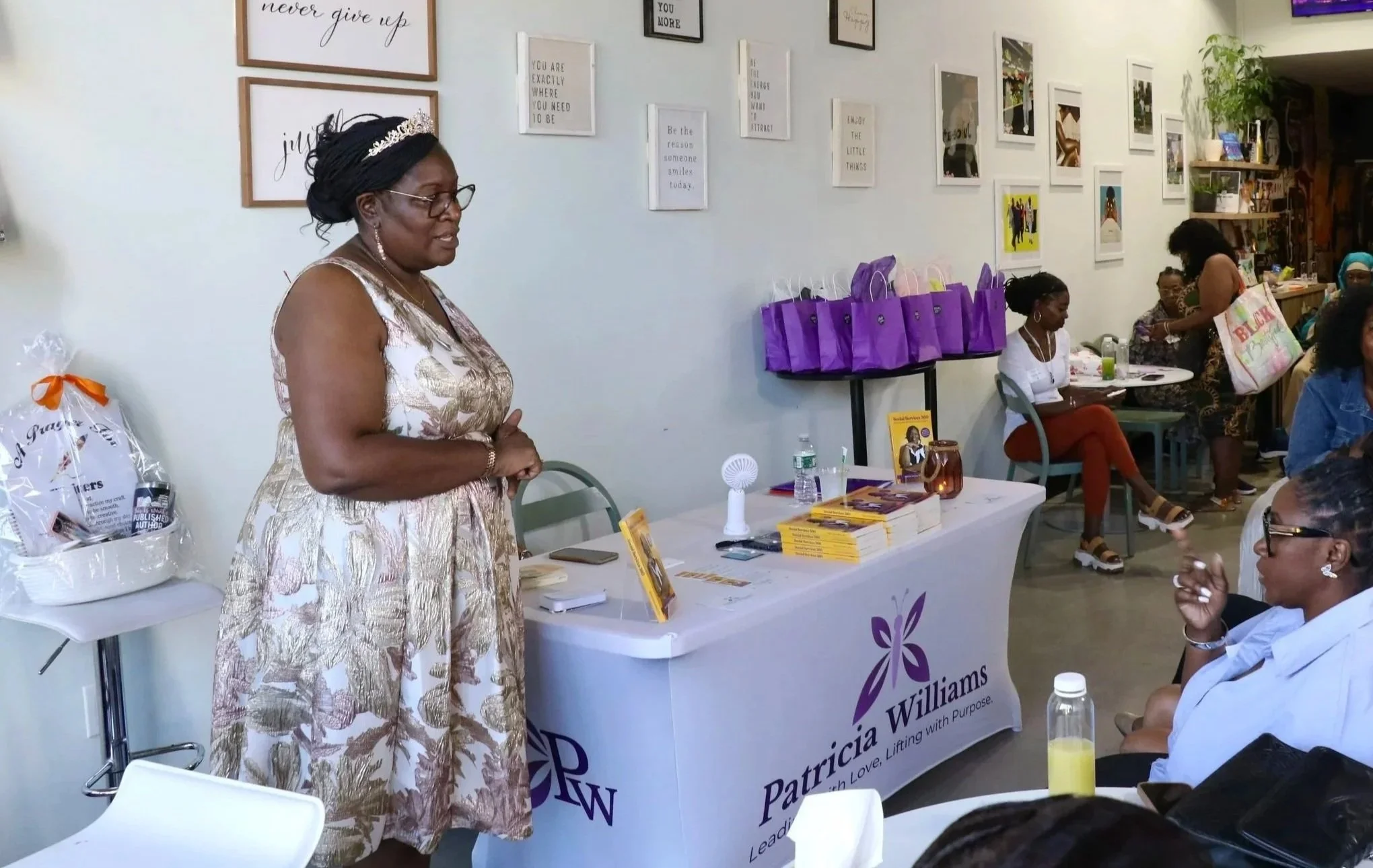 A woman in a floral dress with a tiara and glasses standing in front of a table at a book event, with purple gift bags on the table, displaying books and promotional materials, in a bright space with wall art and several seated women listening.
