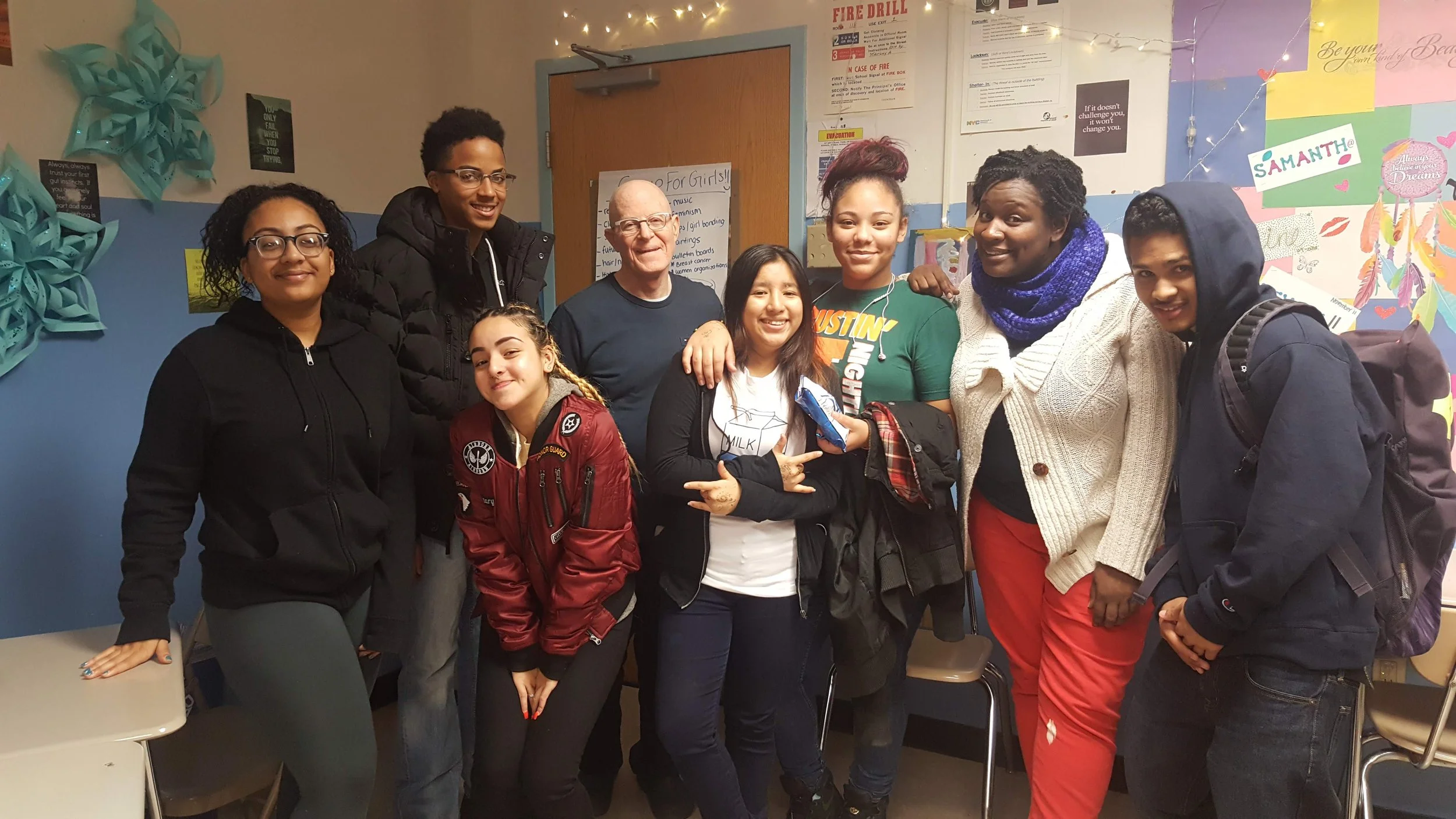 Group of nine diverse people, including children and adults, smiling and posing together inside a room decorated with colorful paper art and posters.