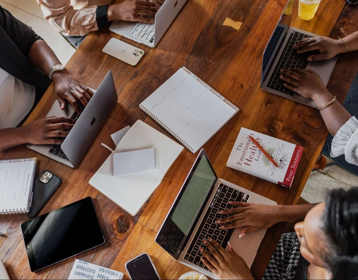 People working on laptops around a wooden table with notebooks, a book titled 'The Emotional Lifestyle' by Peter Scazzero, smartphones, a tablet, a calendar, and drinks.
