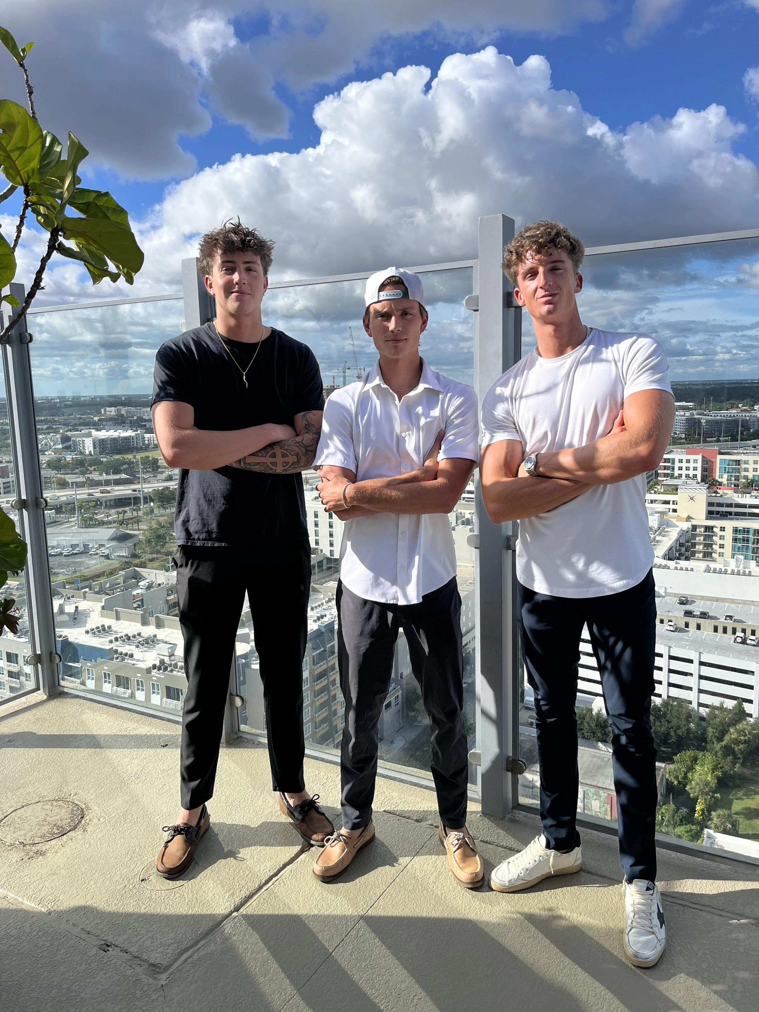 Three young men standing on a rooftop balcony with a cityscape and blue sky with clouds in the background. All have arms crossed and are dressed casually.