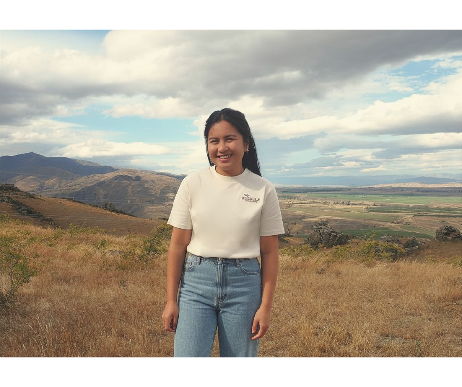 Smiling young girl with dark hair in a ponytail wearing a white t-shirt and denim shorts standing outdoors in a hilly landscape with mountains, farmland, and a partly cloudy sky.