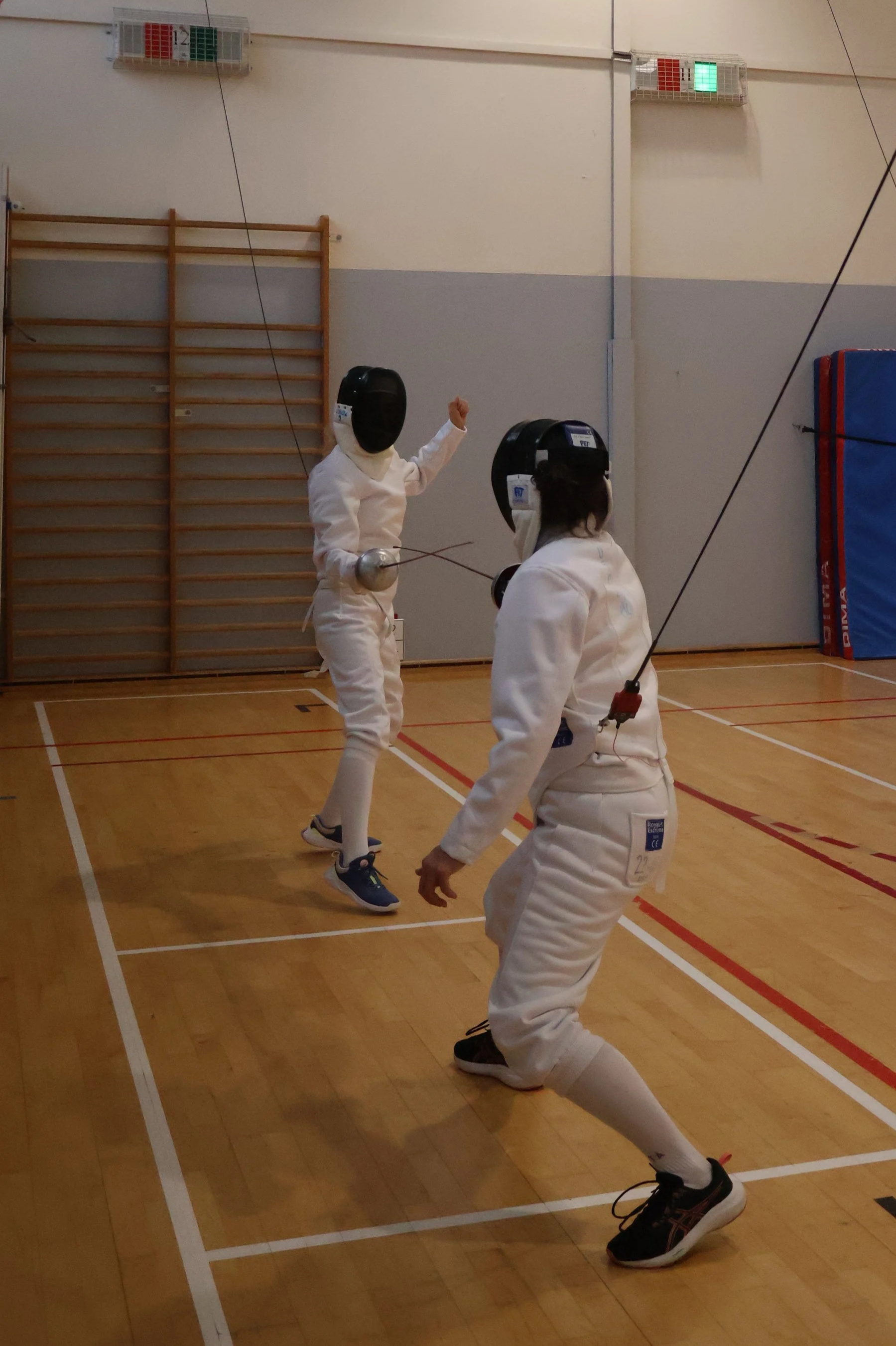 Deux escrimeurs adultes en tenue complète (masque, pantalon d'escrime, veste d'escrime) en cours d'assaut avec des épées branchées à l'électrique dans un gymnase adapté à l'escrime sportive.