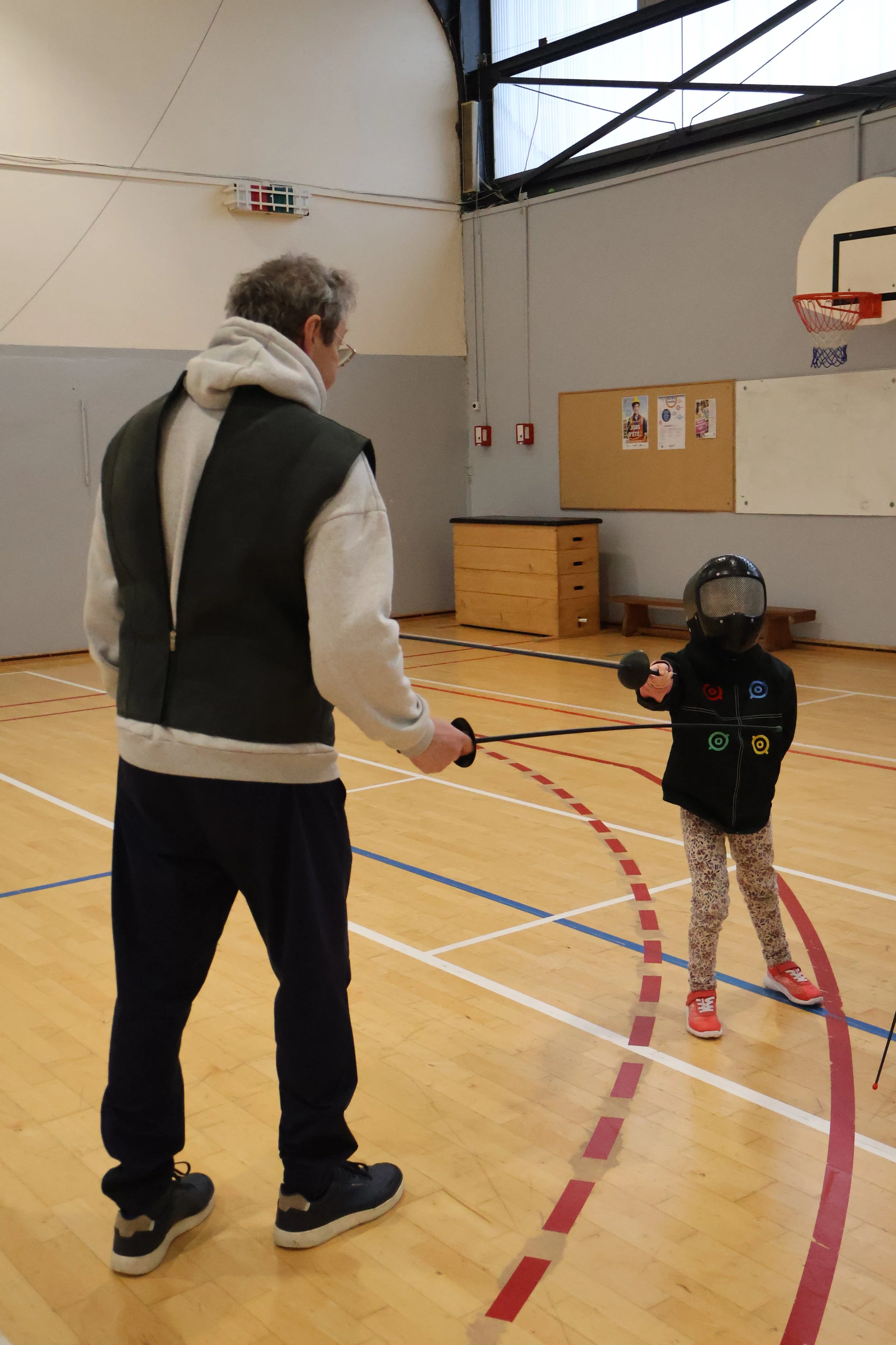 Un maître d'armes et une enfant en équipement d'escrime en train de s'entraîner à la parade.