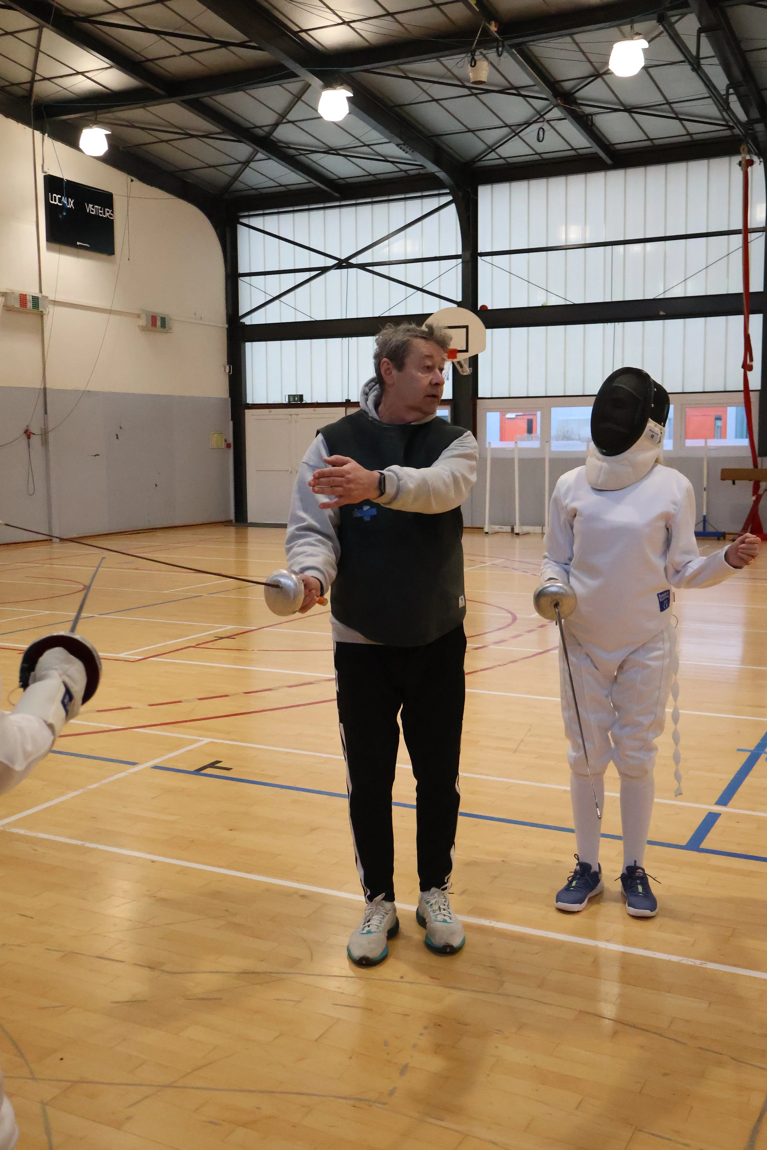 Une personne enseigne la lutte avec épée à deux enfants lors d'une séance pratique dans une salle de sport. Les enfants portent des équipements de protection, notamment un masque intégral.