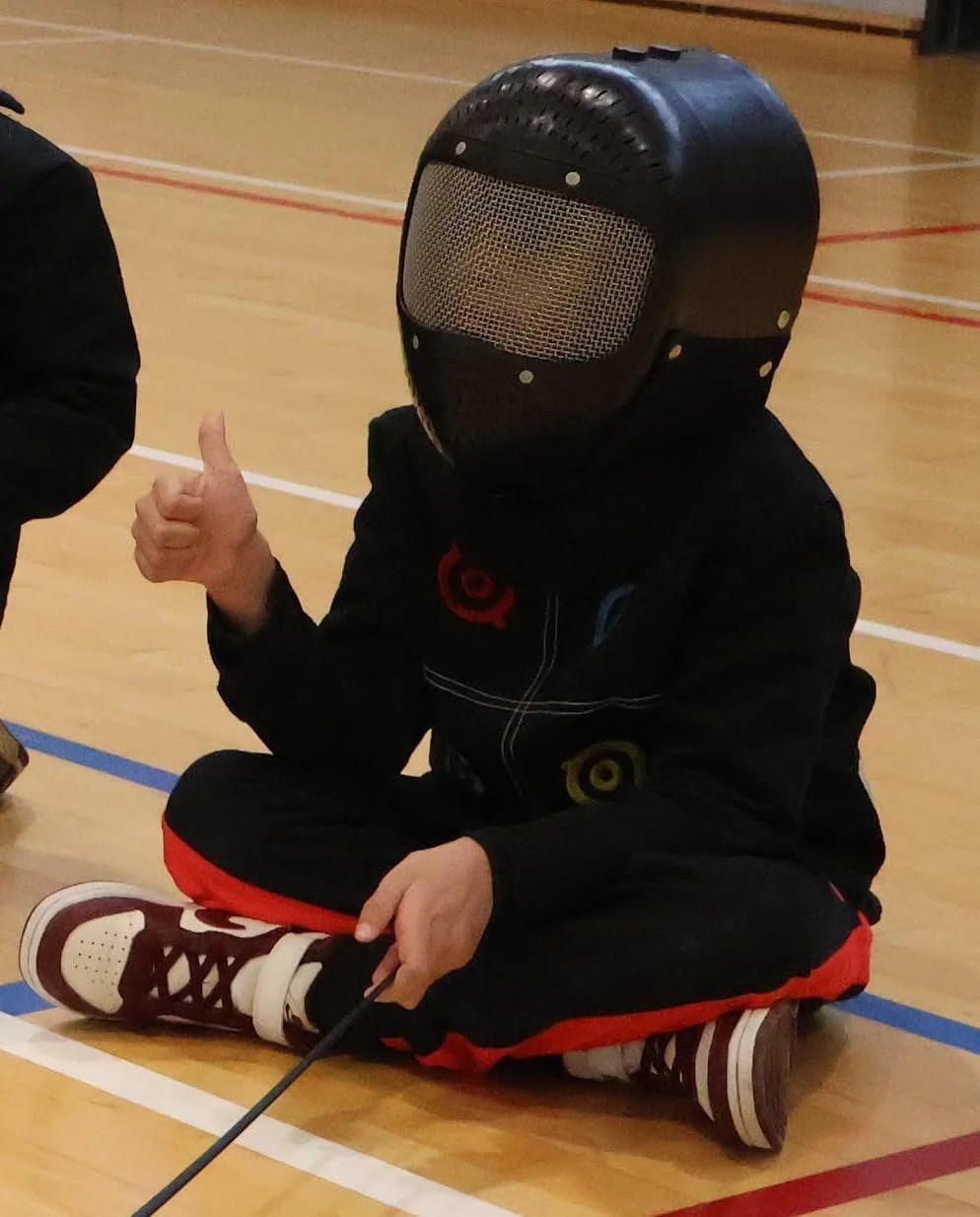 Jeune enfant en tenue d'escrime, épée, fleuret ou sabre, assise par terre, faisant un signe de pouce en l'air, dans une salle d'escrime.