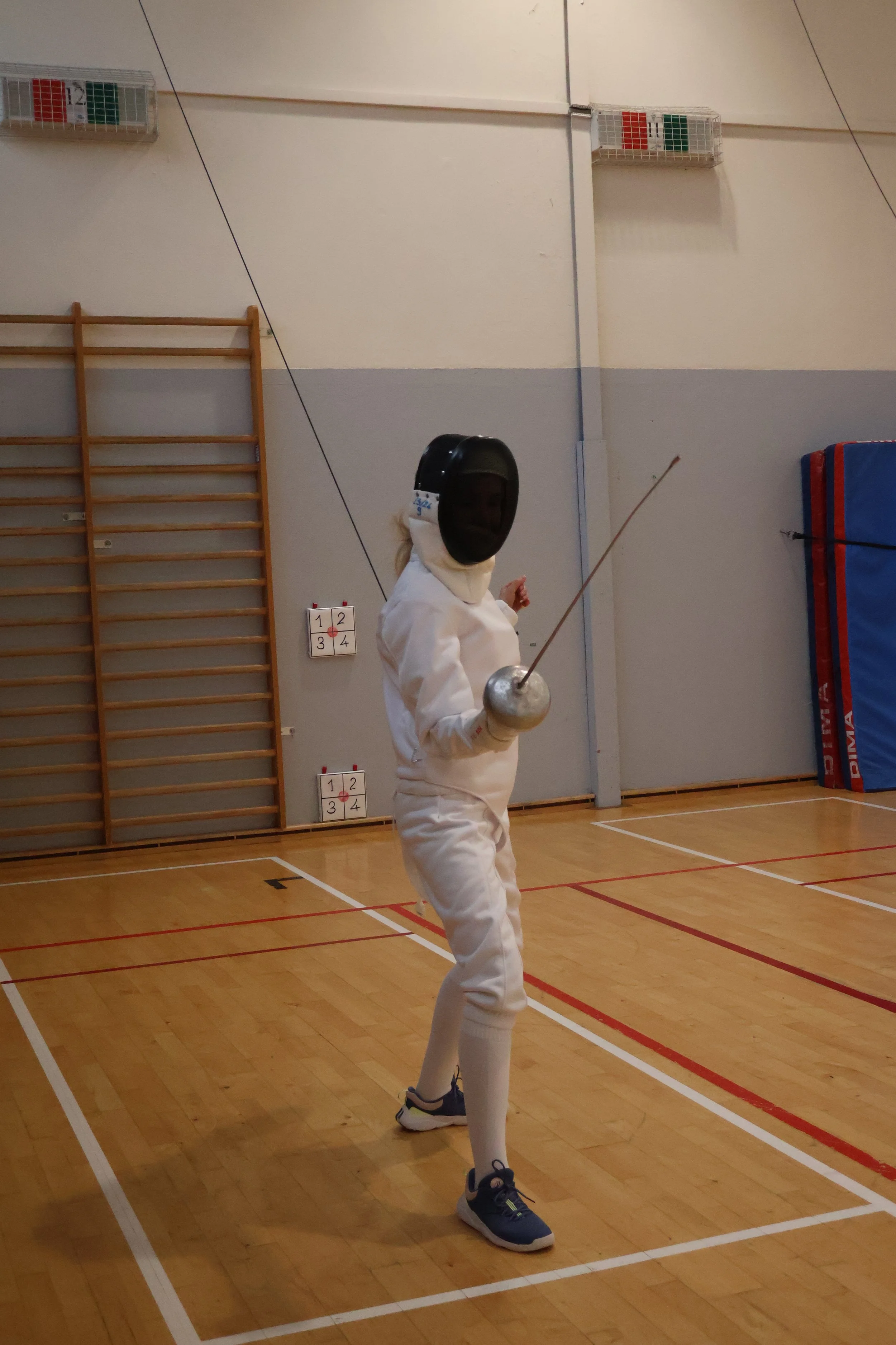 Un(e) escrimeur(se) en tenue d'escrime avec masque, veste d'escrime, pantalon d'escrime, tenant une épée dans une salle d'escrime