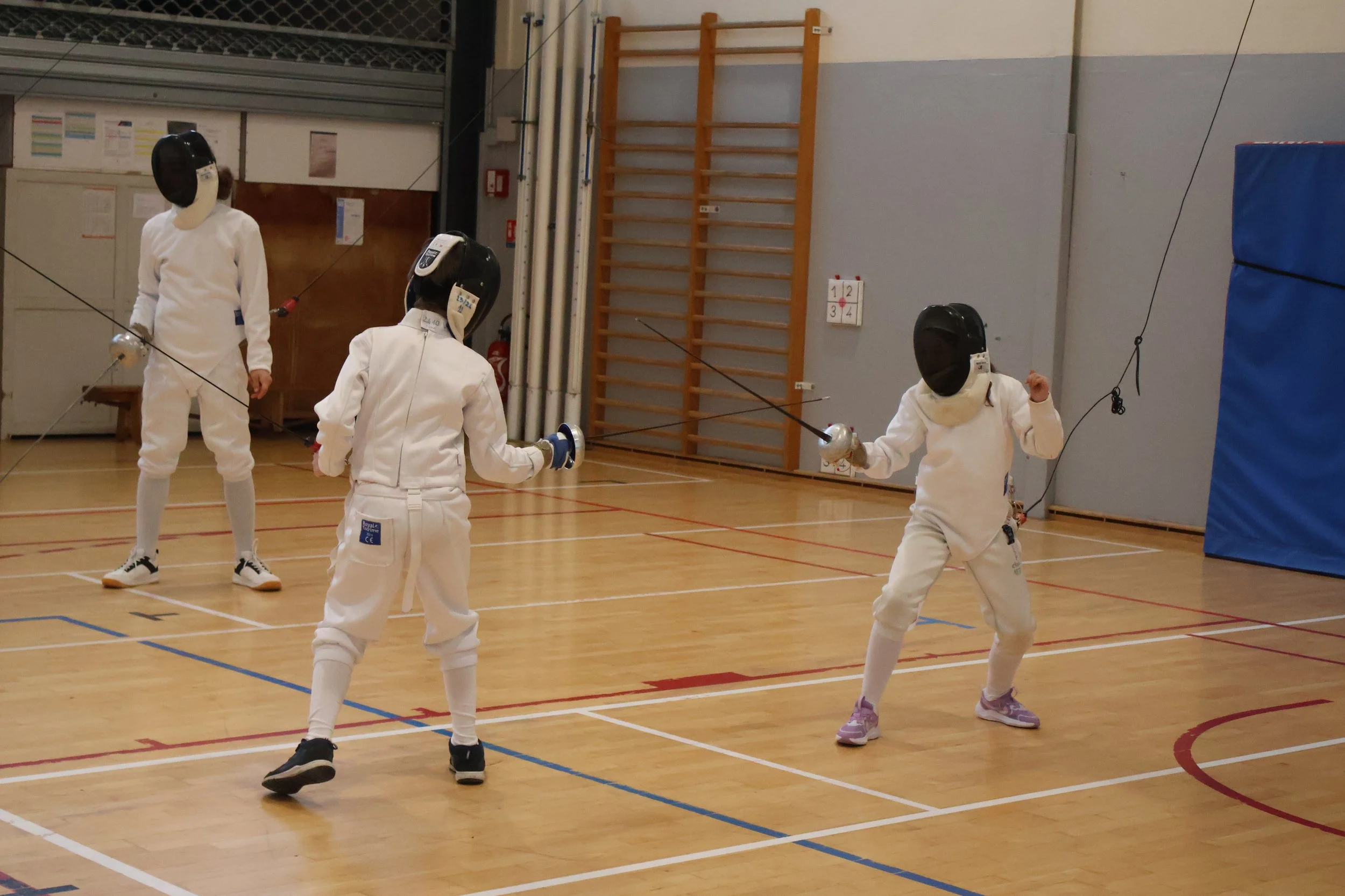 Quatre jeunes en équipement d'escrime, pratiquant l'escrime en intérieur.