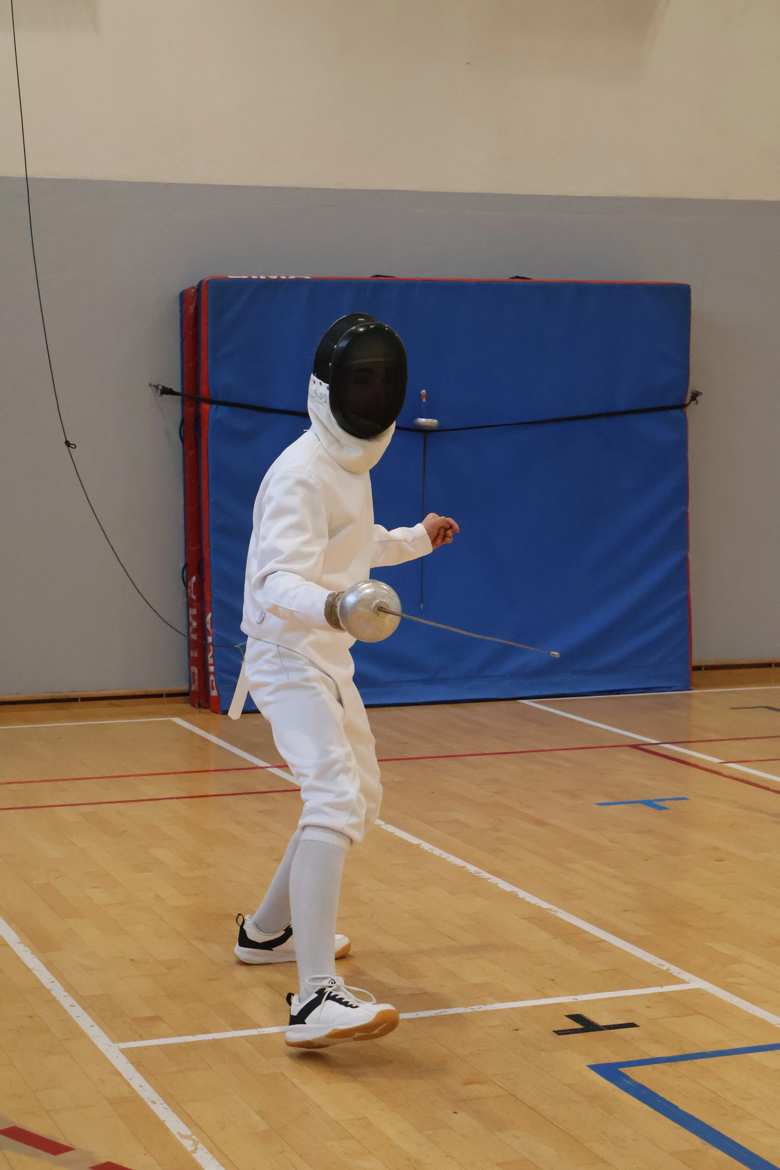 Jeune tireur en tenue blanche et masque de protection, tenant une épée, pratiquant l'escrime dans une salle d'armes.