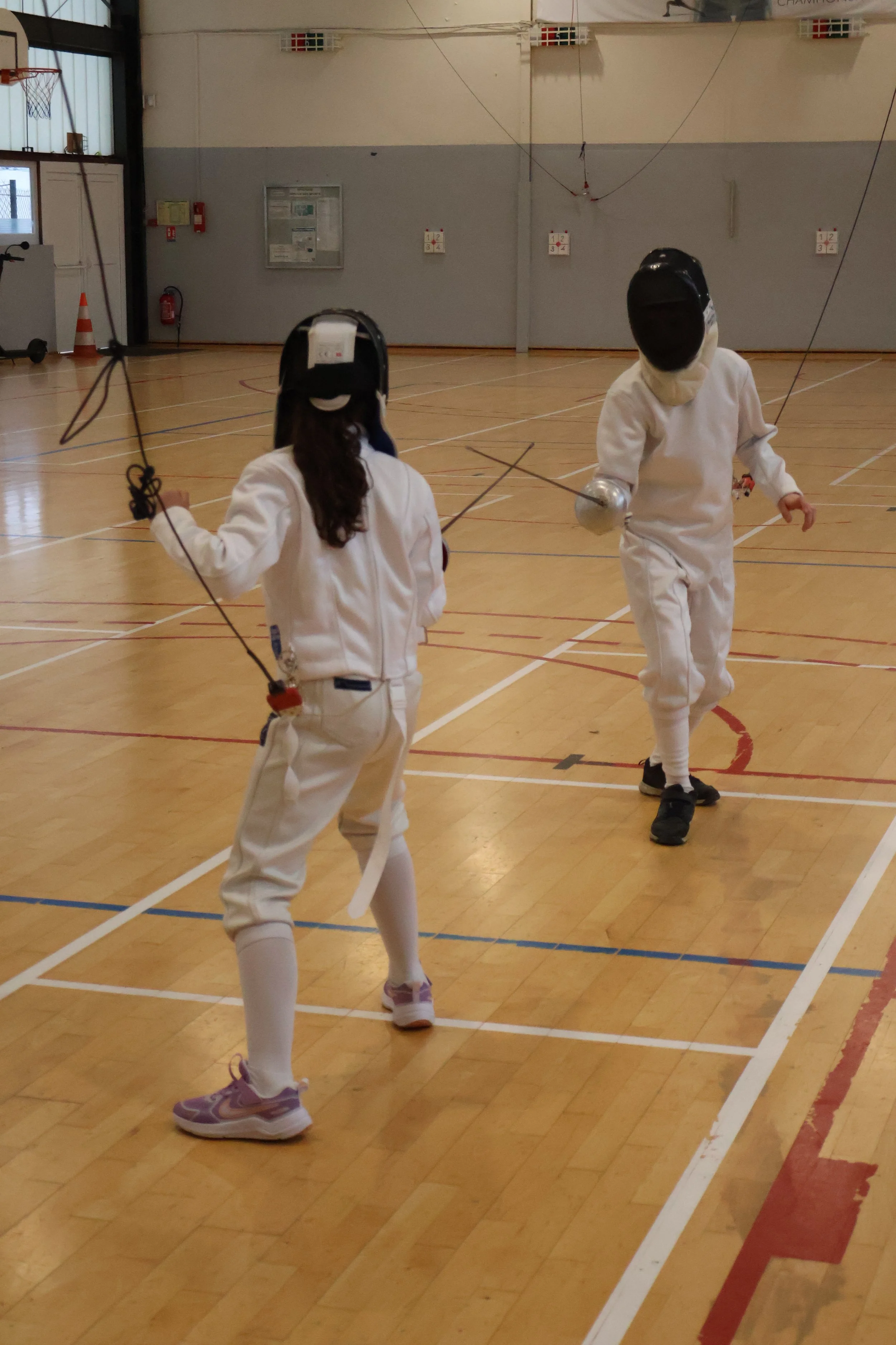 Deux enfants pratiquant l'escrime dans une salle de sport, équipés de tenues d'escrime et de masques de protection