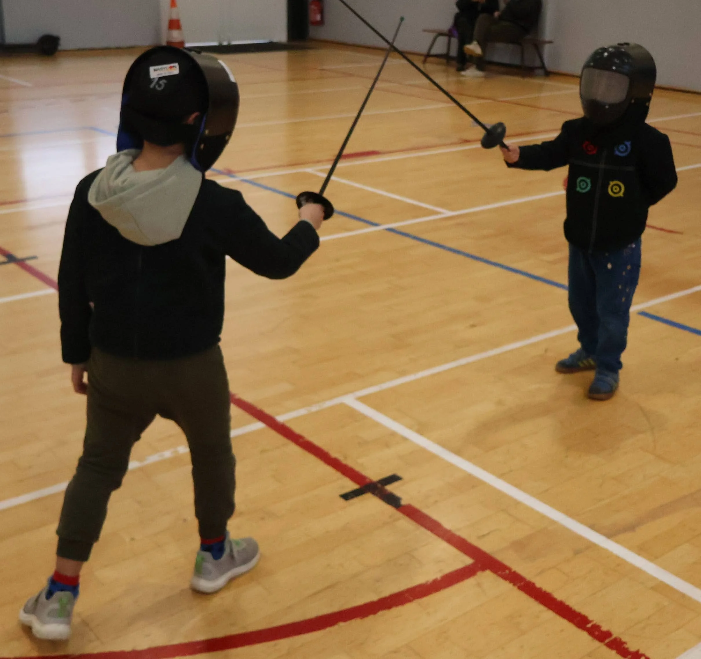Deux enfants entre 4 et 7 ans lors d'un duel à l'épée en plastique dans une salle d'estime, portant un masque et une veste d'escrime
