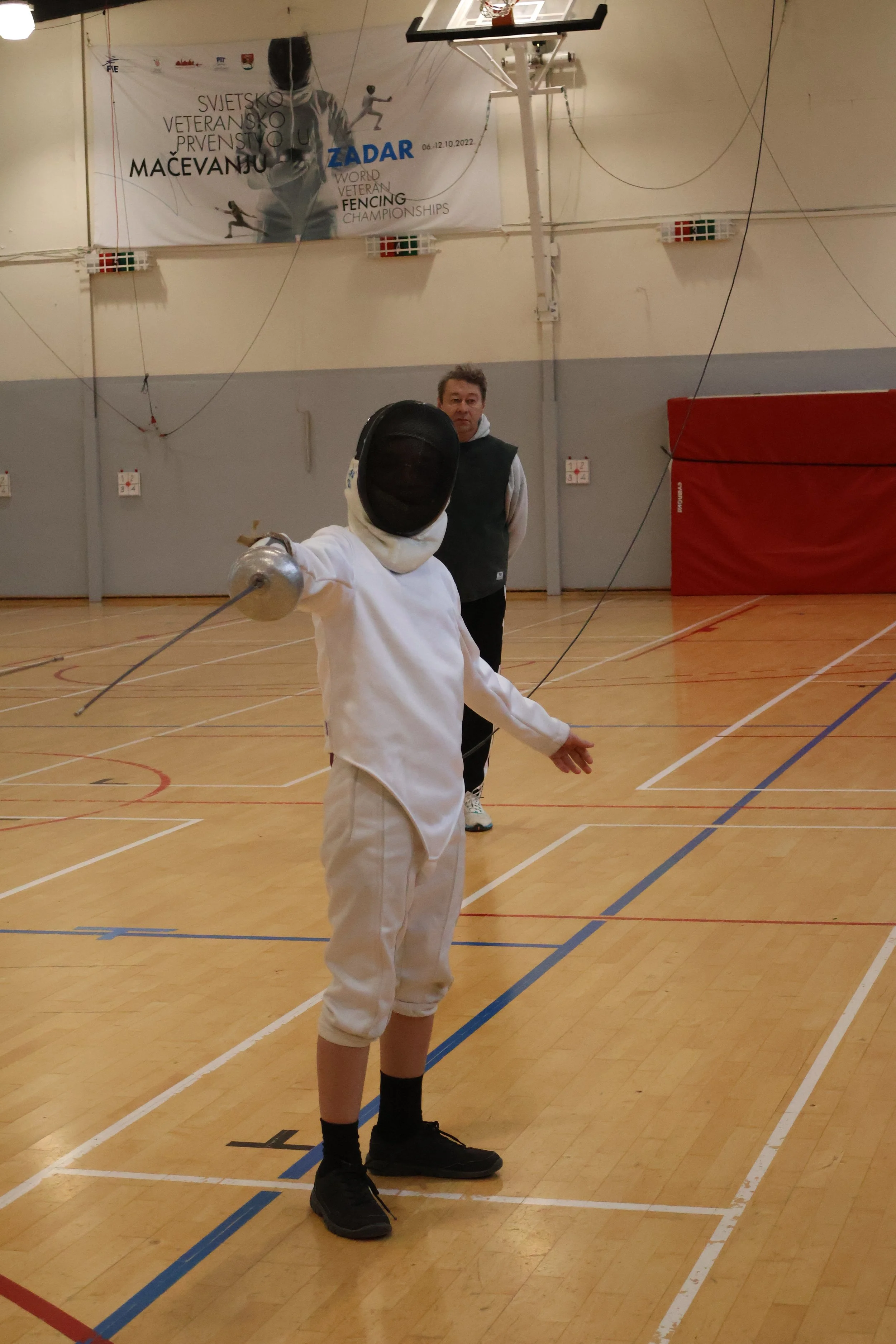 Un escrimeur adolescent lors d'une séance d'apprentissage de l'escrime, en position d'attaque bras tendu, avec un maître d'arme qui observe en arrière-plan.
