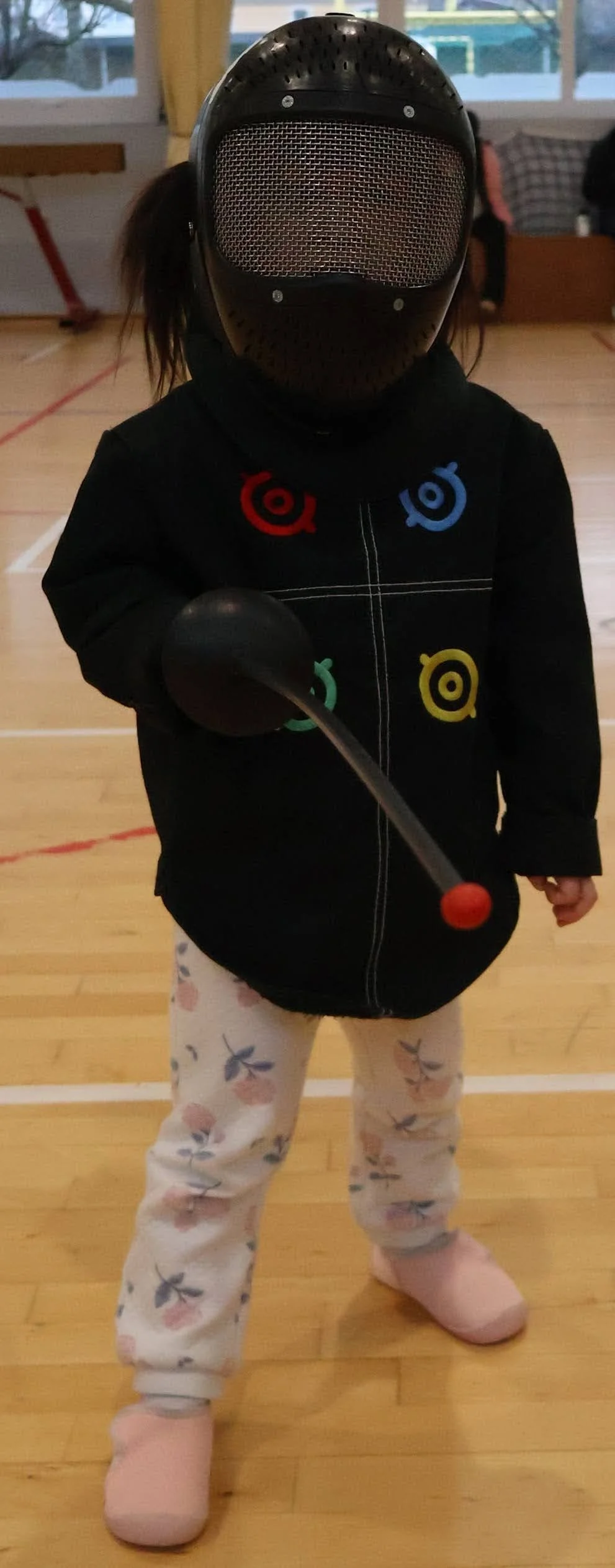 Enfant à partir de 4 ans en tenue d'escrime portant un masque d'escrime, une veste d'escrime et tenant un fleuret en plastique, dans un gymnase adapté à la pratique des l'escrime sportive.