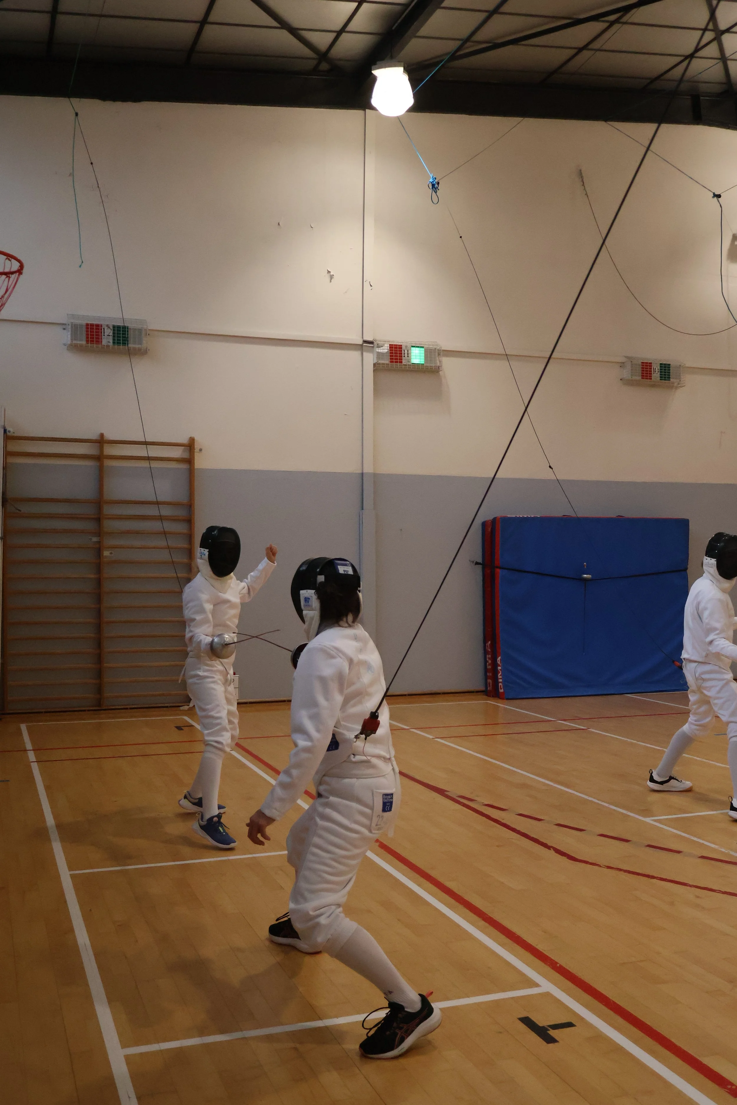 Deux personnes pratiquant l'escrime dans une salle d'escrime, équipées dune tenue d'escrime complète avec masques d'escrime, veste d'escrime et pantalon d'escrime, tenant des épées.