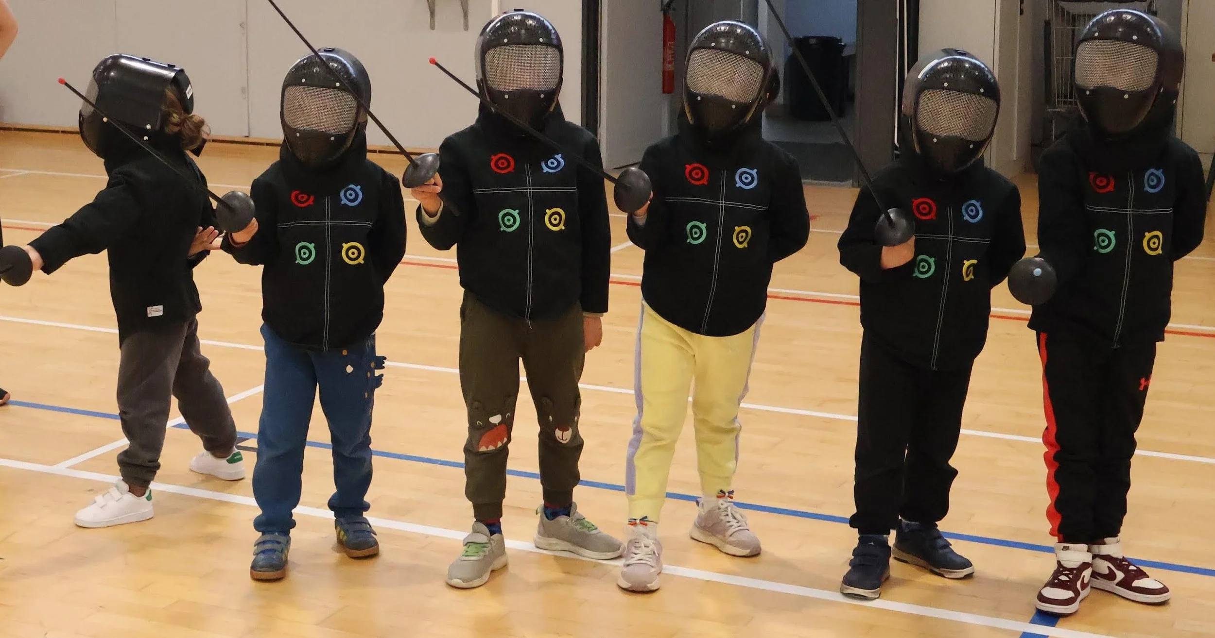 Six enfants entre 4 et 7 ans en tenue d'escrime avec des masques et des épées en plastique, se tenant en position de garde sur une piste d'escrime.