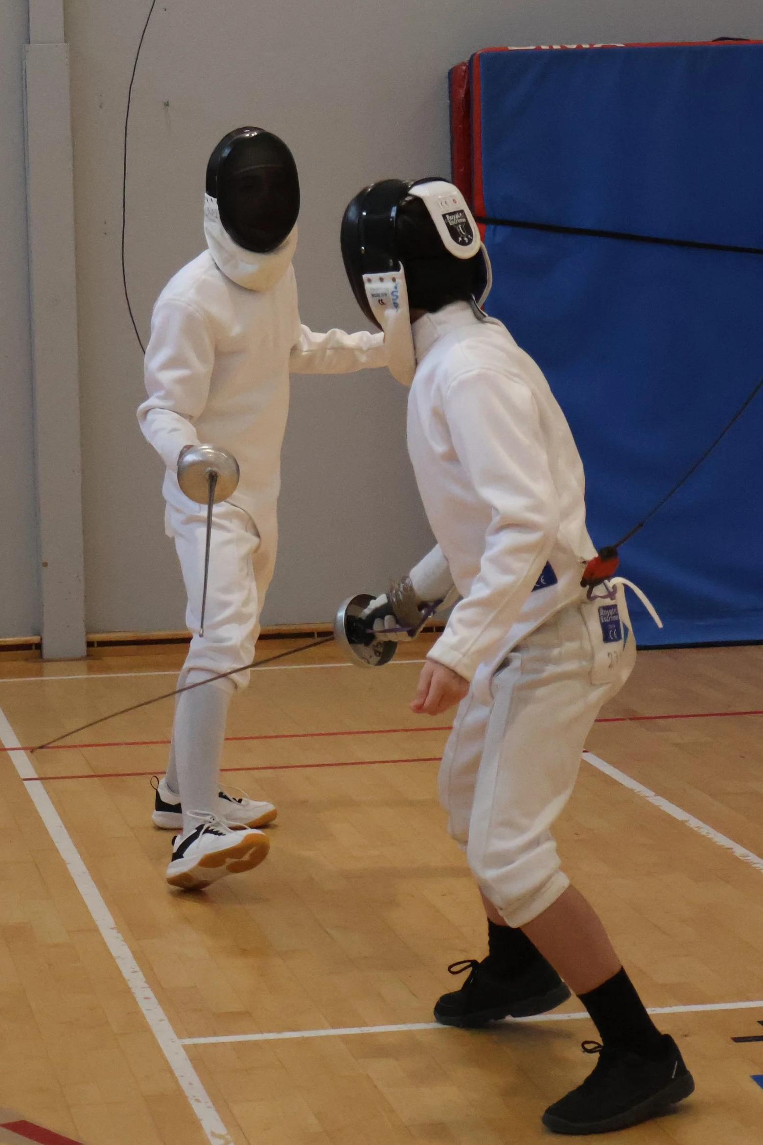 Deux escrimeurs adolescents pratiquant l'escrime en salle, équipées de vêtements de protection et de masques, en train de s'affronter à l'épée électrique.