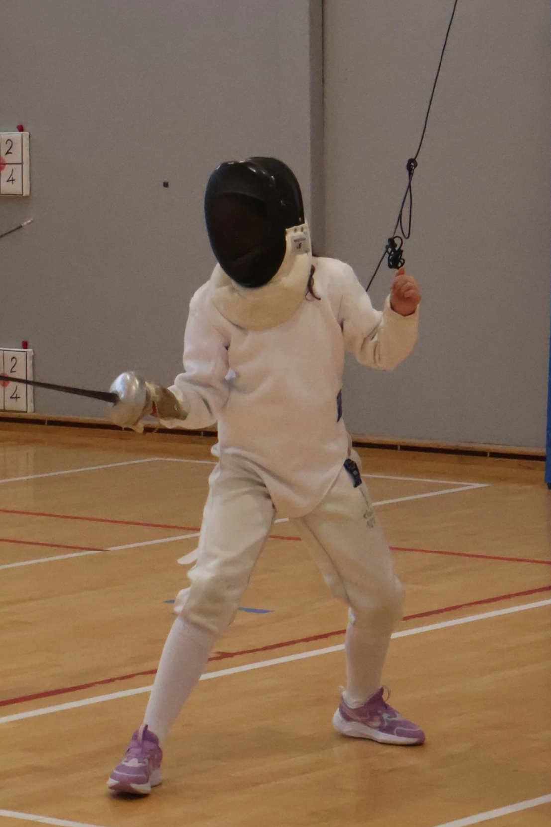 Une jeune enfant en position de garde dans une salle d'escrime, tenant une épée et portant une tenue complètes d'escrime (un masque d'escrime, pantalon d'escrime, veste d'escrime).