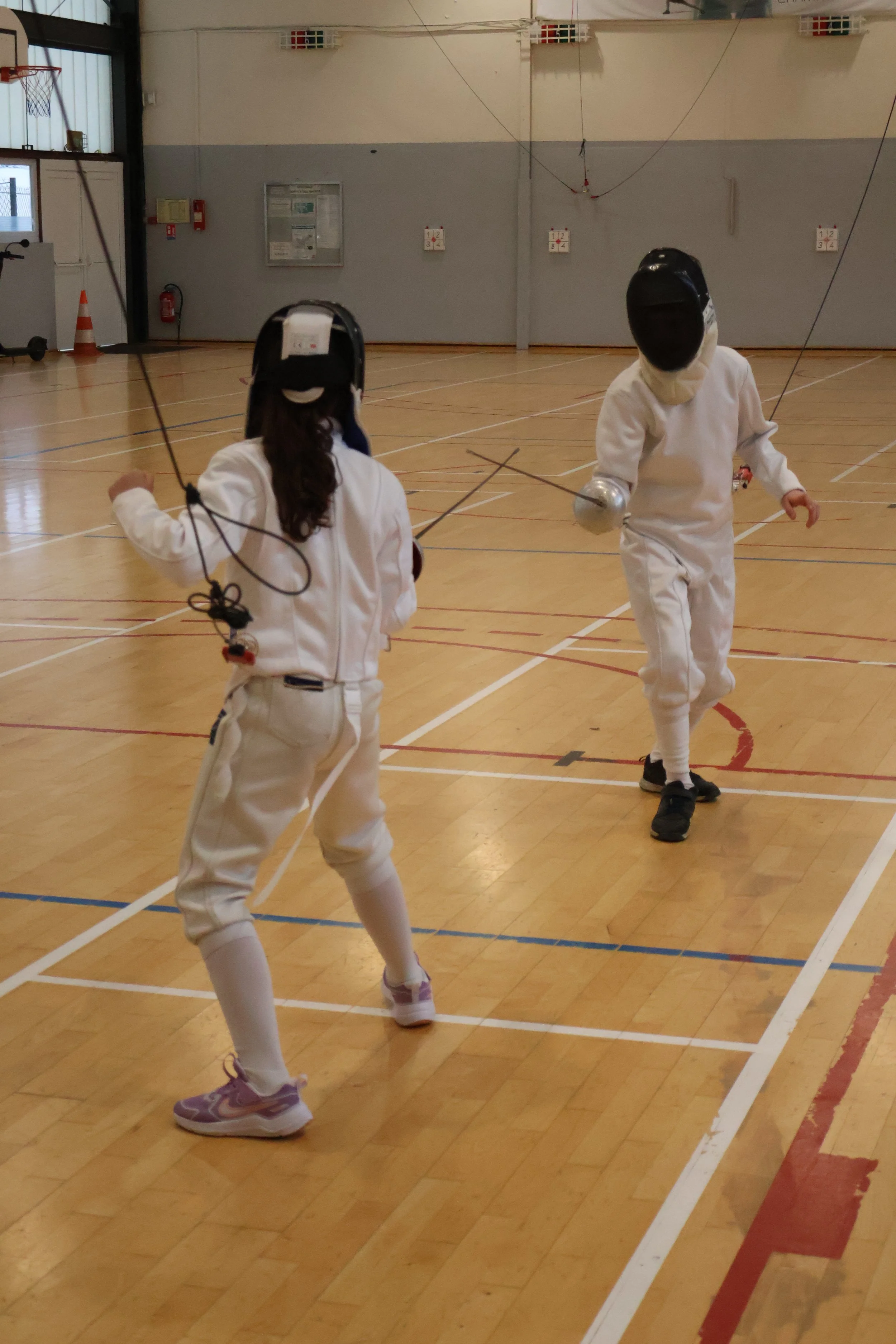 Deux tireurs enfants, une fille face à un garçon, en tenue d'escrime  (masque, pantalon d'escrime, veste d'escrime)  s'entraînent dans un gymnase lors d'un duel avec des épées branchées à l'électrique.