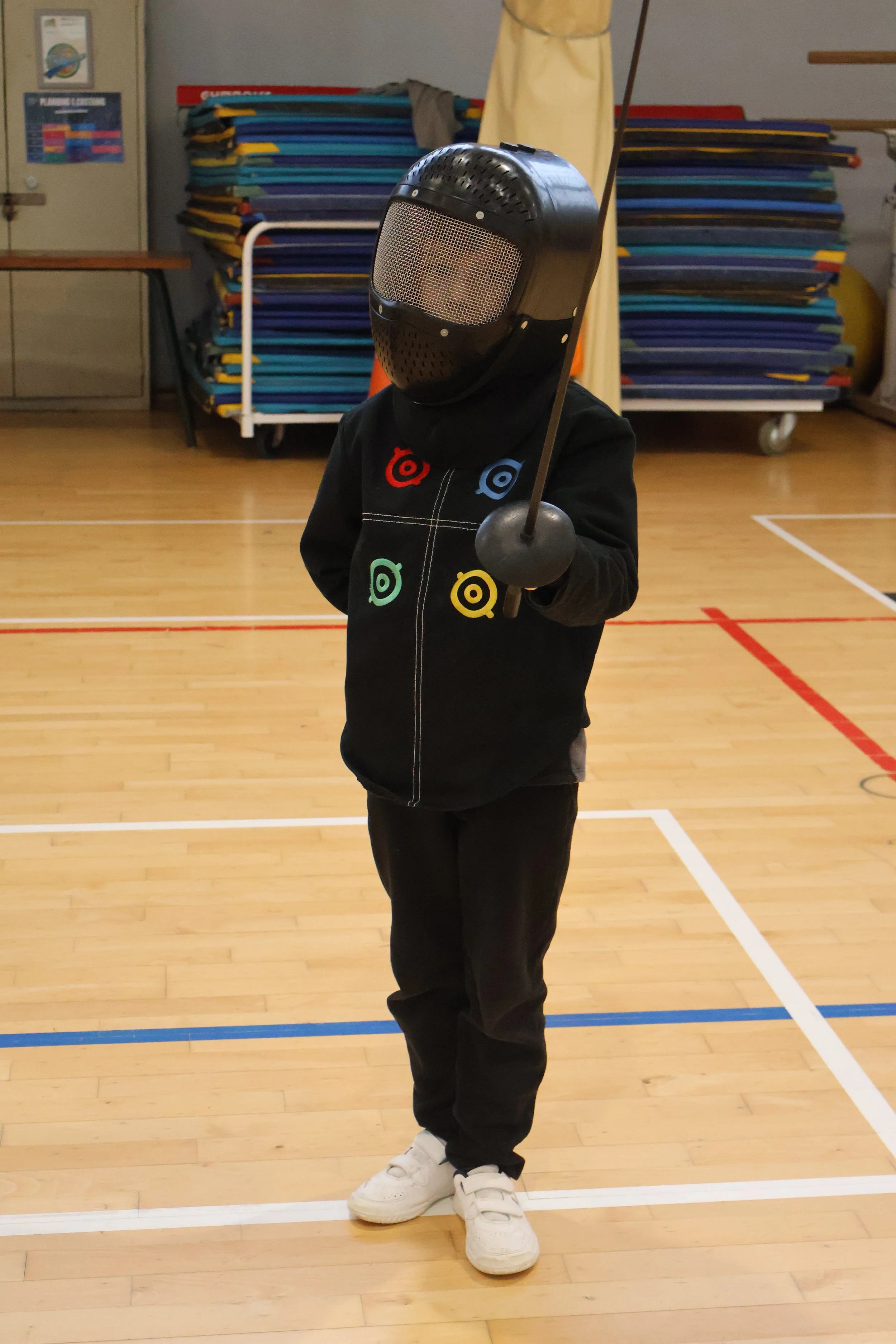 Enfant en tenue costume de escrime avec masque et arme, dans une salle de sport.