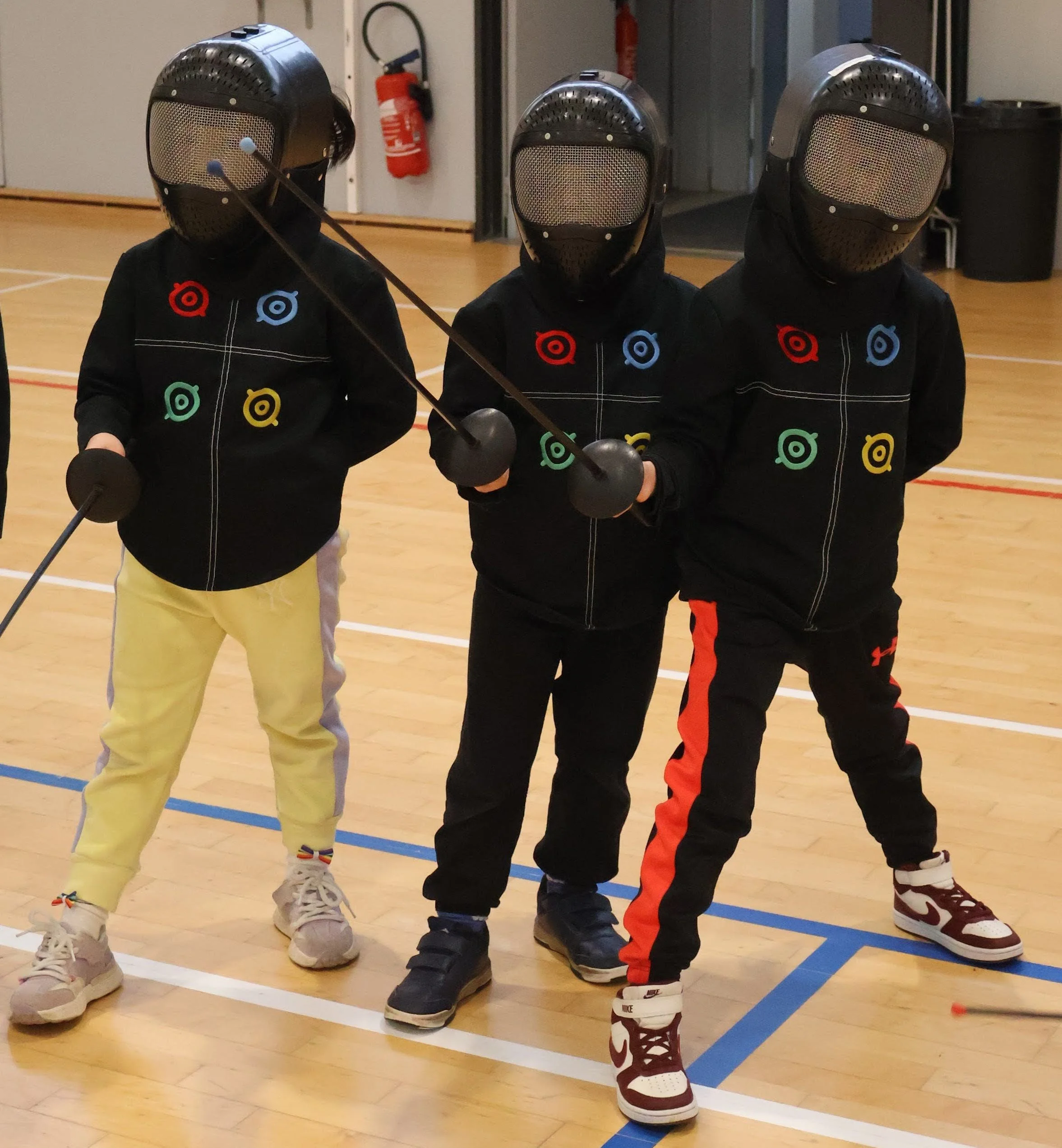 Trois enfants en vêtements de sport portant des masques pour la pratique de l'escrime, tenant des épées.