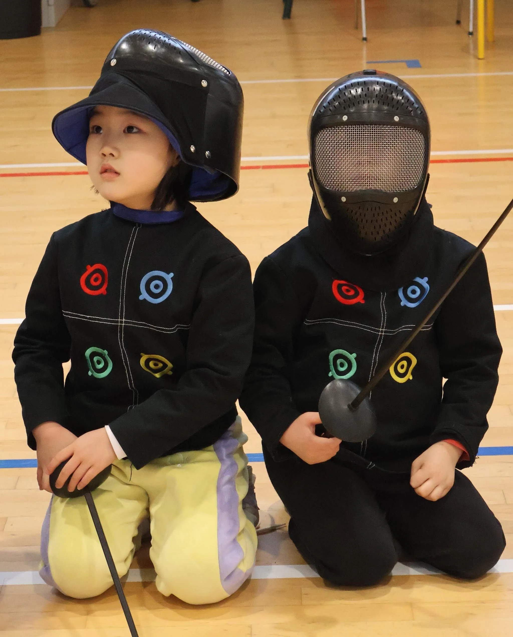 Deux enfants en tenue d'escrime, portant des masques, à l'écoute du maître d'arme.