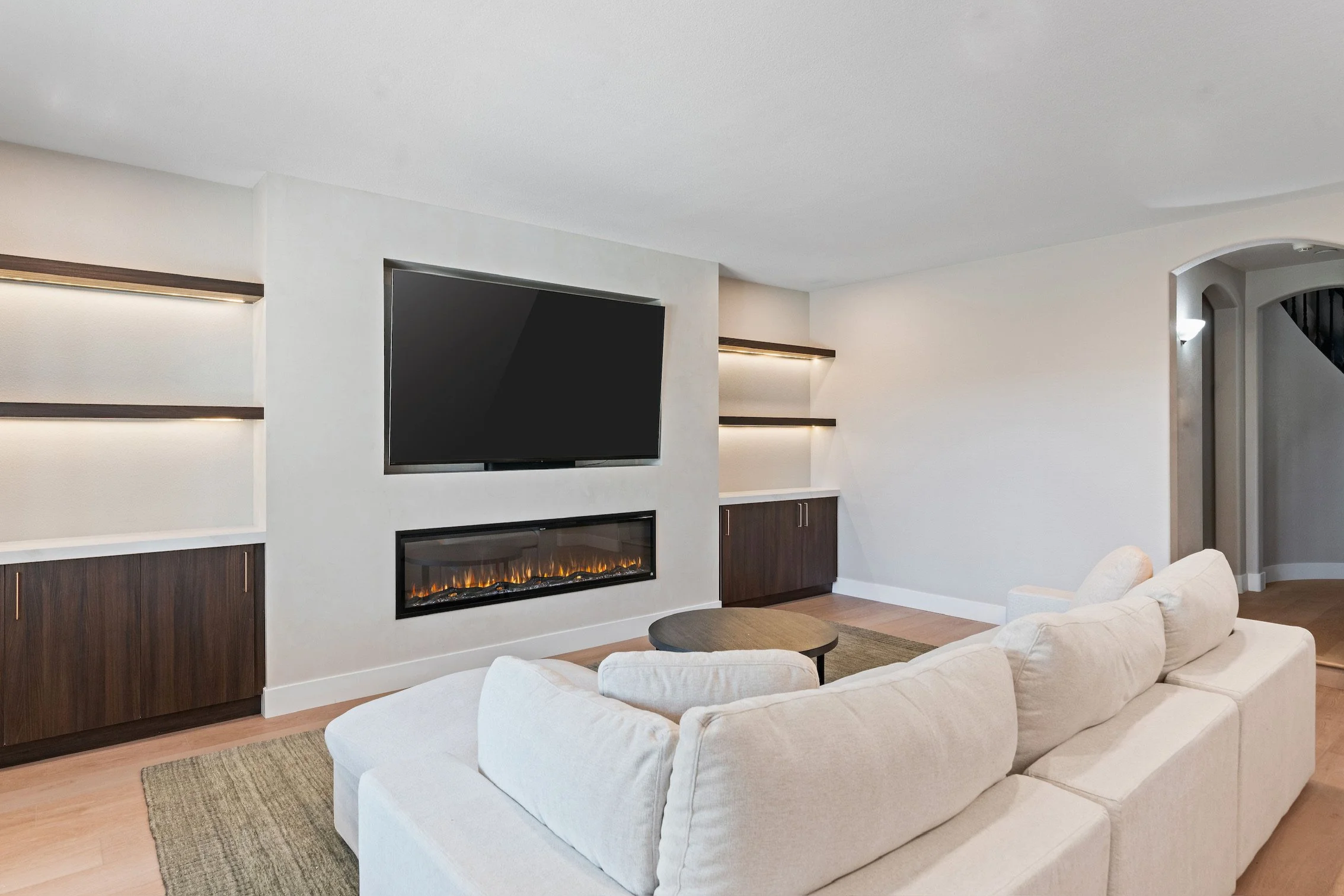 Living room with a beige sofa, a wall-mounted TV, and a modern fireplace below the TV, with built-in shelves and cabinets on either side.