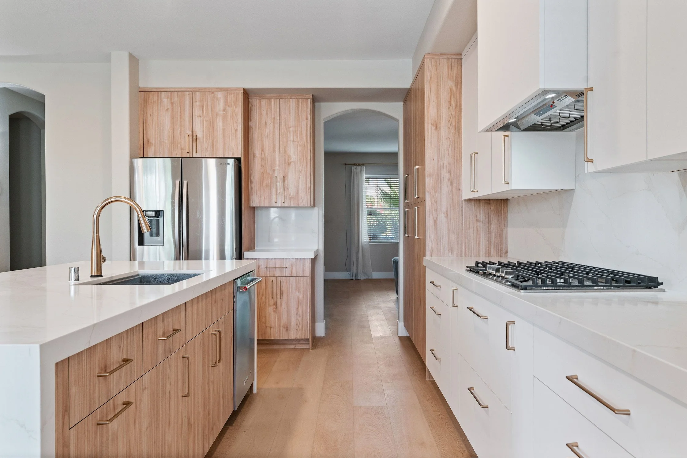 Modern kitchen with white and wood cabinets, a stainless steel refrigerator, a gas stove, a white countertop with a sink, and a view into a dining area with a window and curtains.