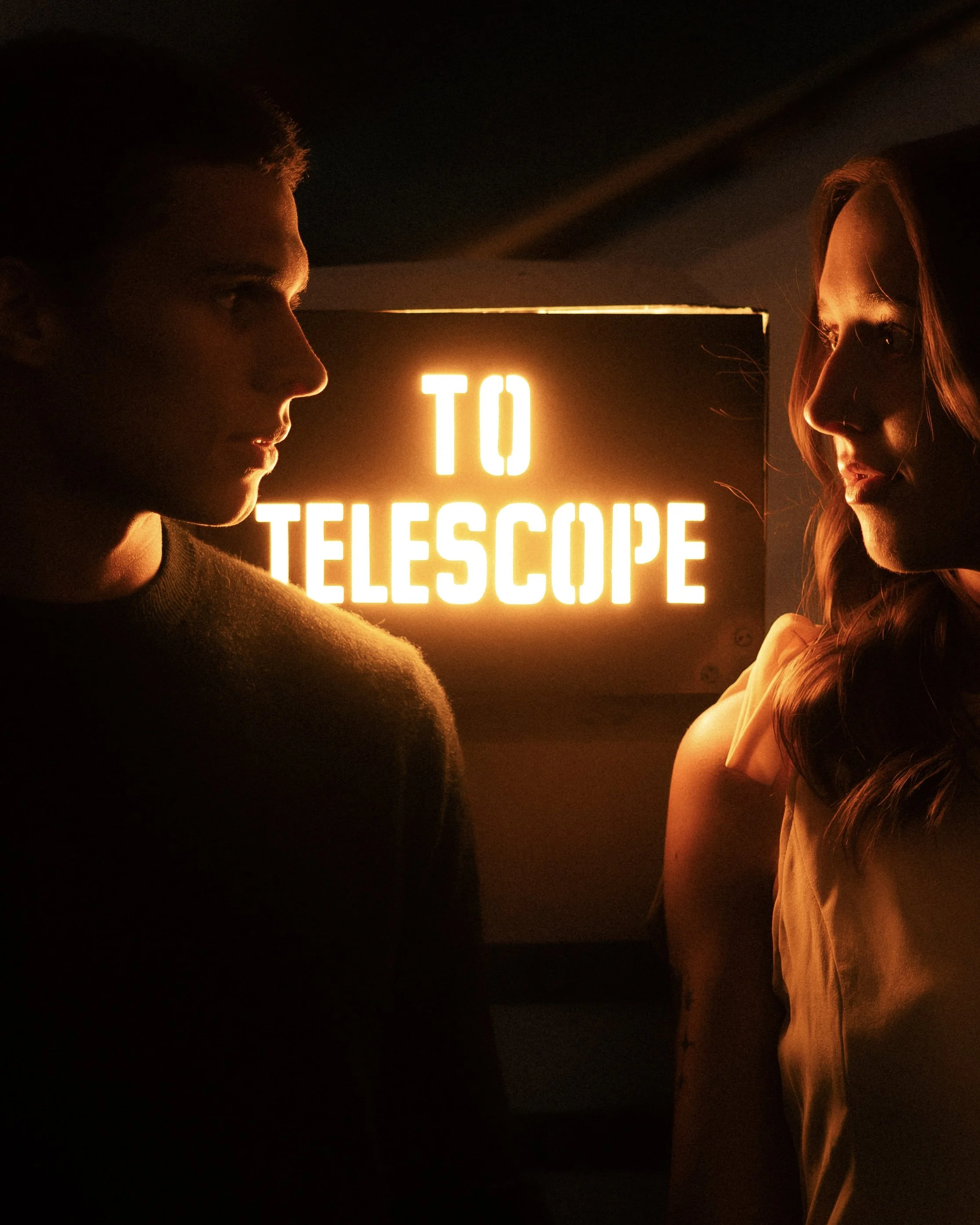 Two people facing each other in a dark room with a bright sign that reads 'TO TELESCOPES' behind them.