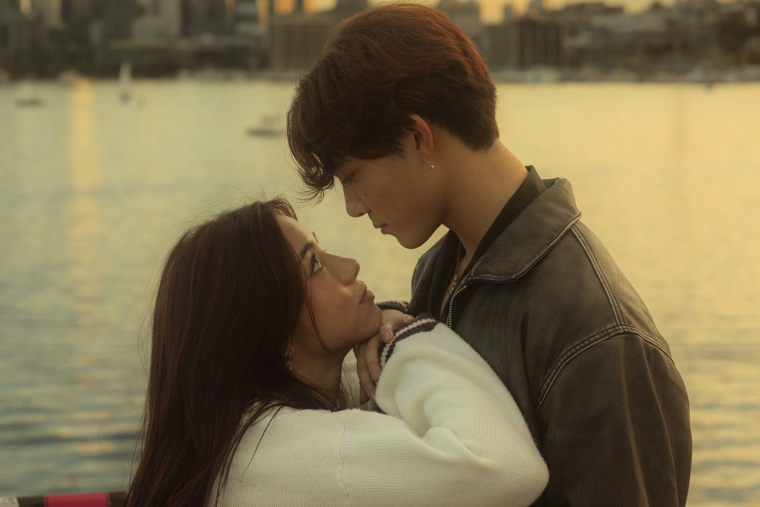 A man and woman are embracing by a body of water during sunset, with the man's forehead gently touching the woman's forehead.