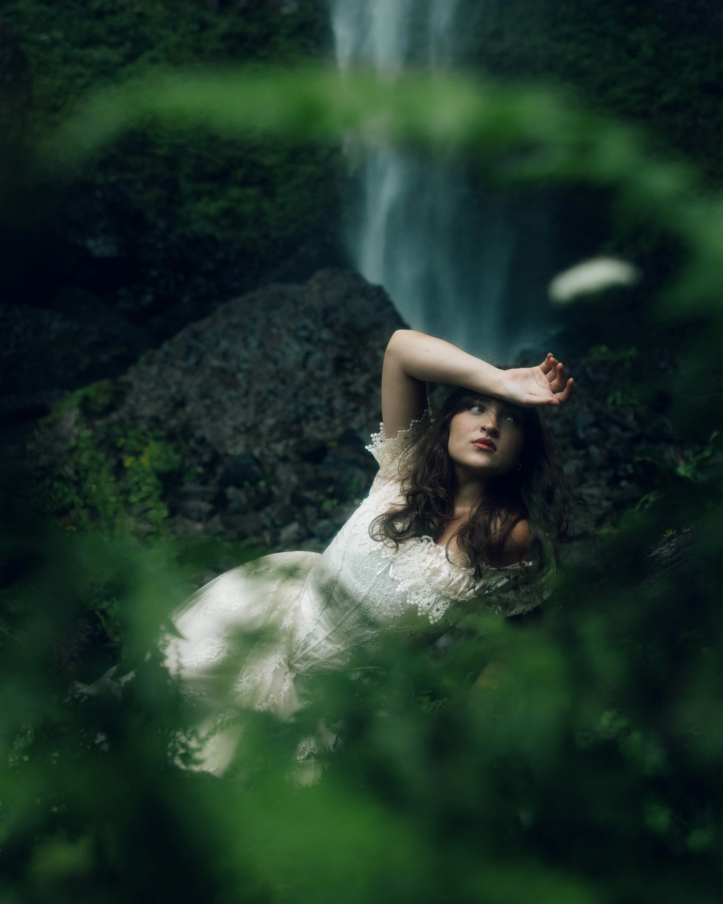 A woman in a white dress lying on the ground among rocks and green foliage near a small waterfall in a forest, with her arm resting above her head.