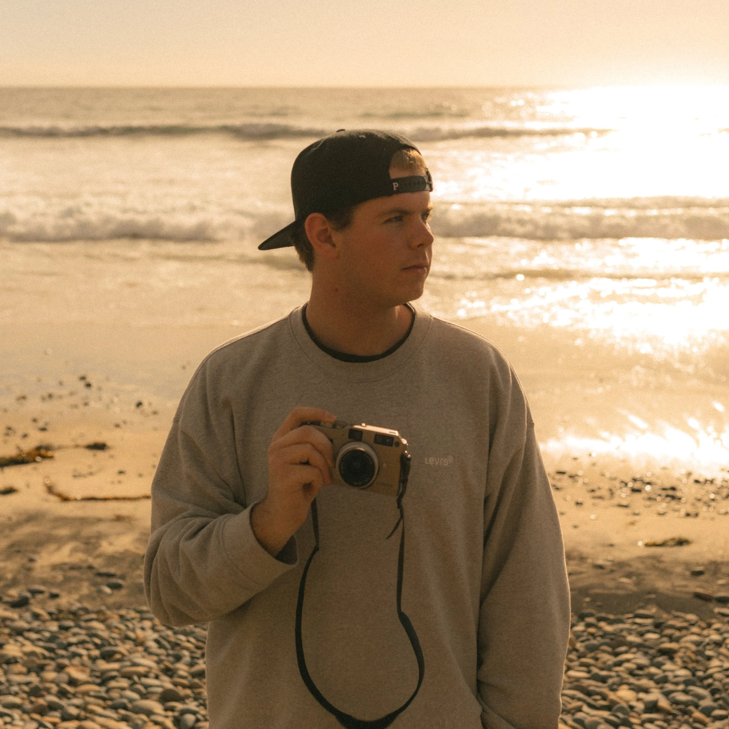 A young man in a gray Levi's sweatshirt and backwards black baseball cap standing on a pebbled beach at sunset, holding a camera with the ocean and a bright sunlight reflection in the background.